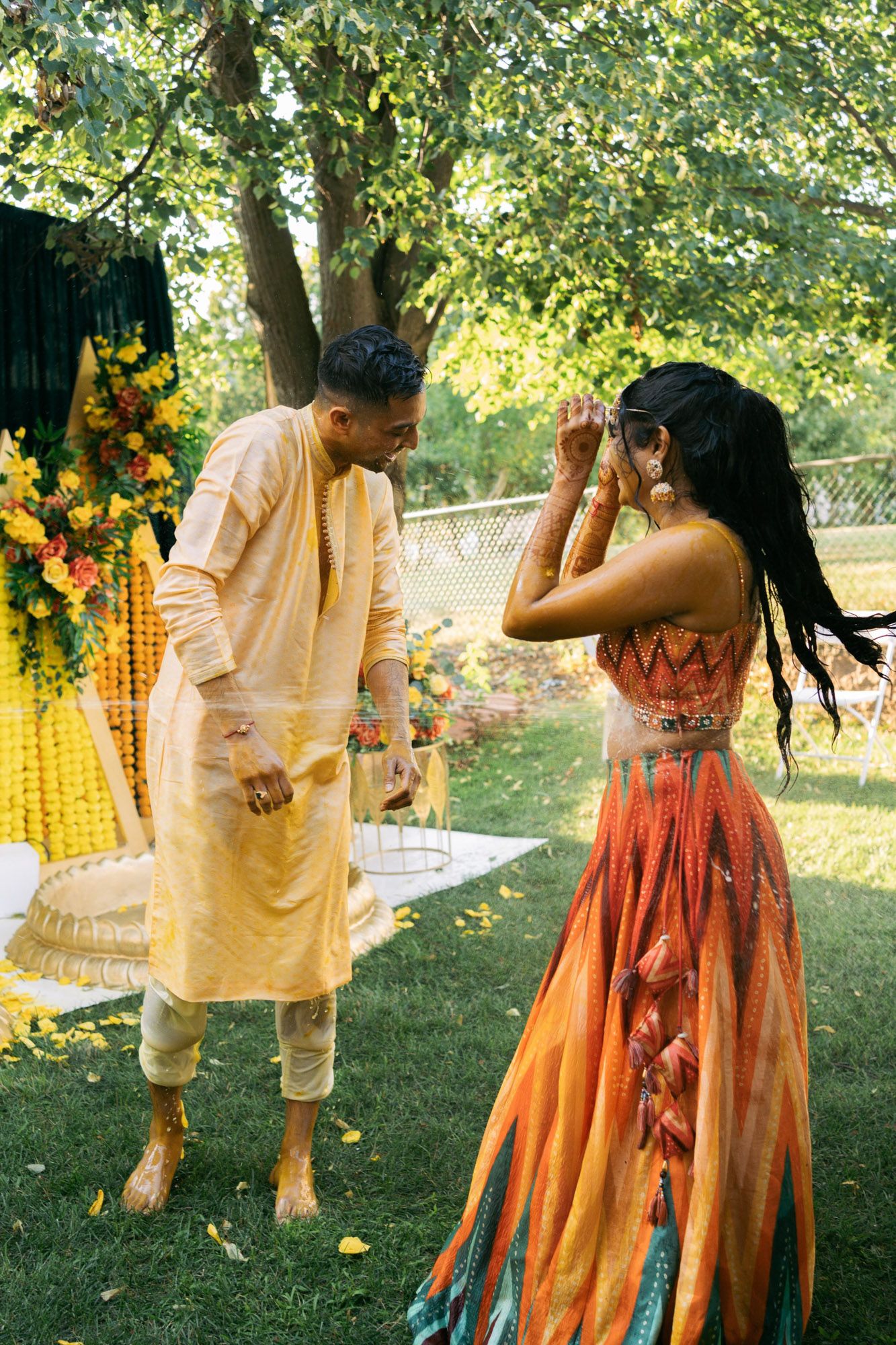 A couple in traditional Indian attire dances together in a sunlit garden setting with yellow and orange decorations.