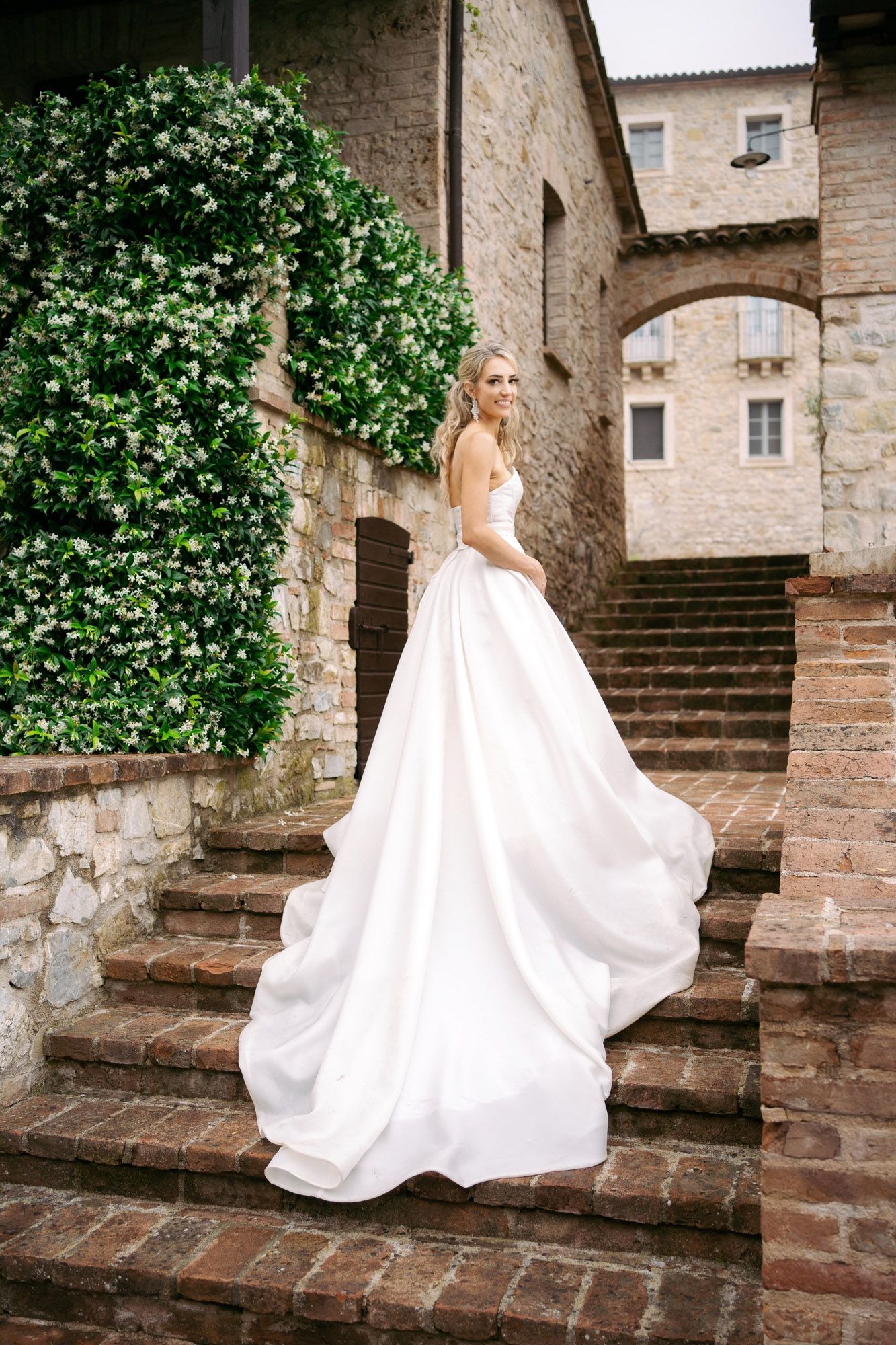 A figure in a flowing white wedding dress poses gracefully on rustic brick stairs against an old stone wall.