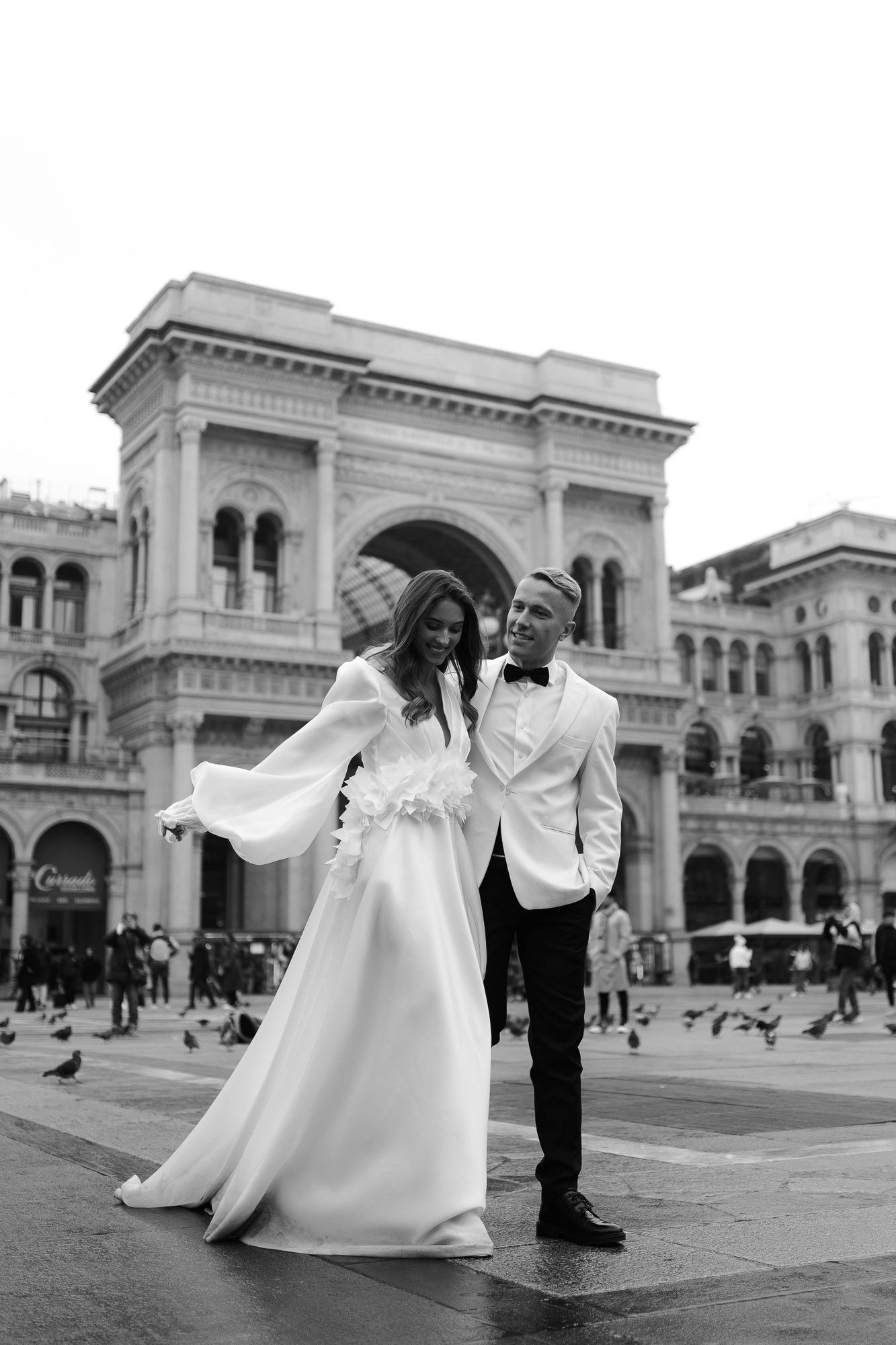 A couple in wedding attire dances together in front of an elegant classical building with arched architecture.