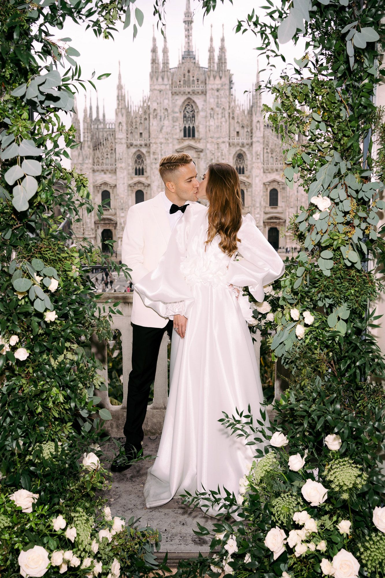 Romantic wedding couple embraces on a terrace overlooking the historic Milan Cathedral with white roses and greenery framing them.