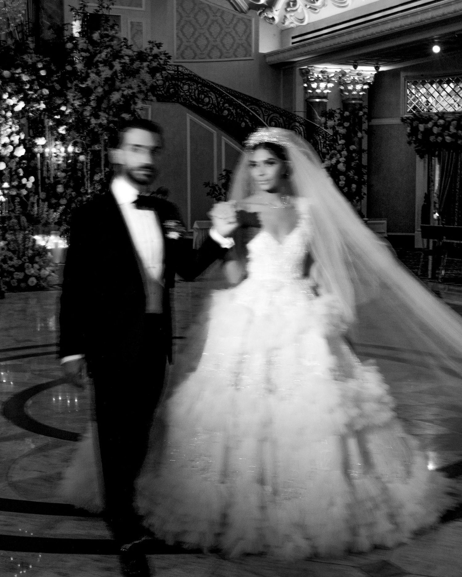 A black and white photograph of a bride and groom walking together in an elegant ballroom with floral decorations.