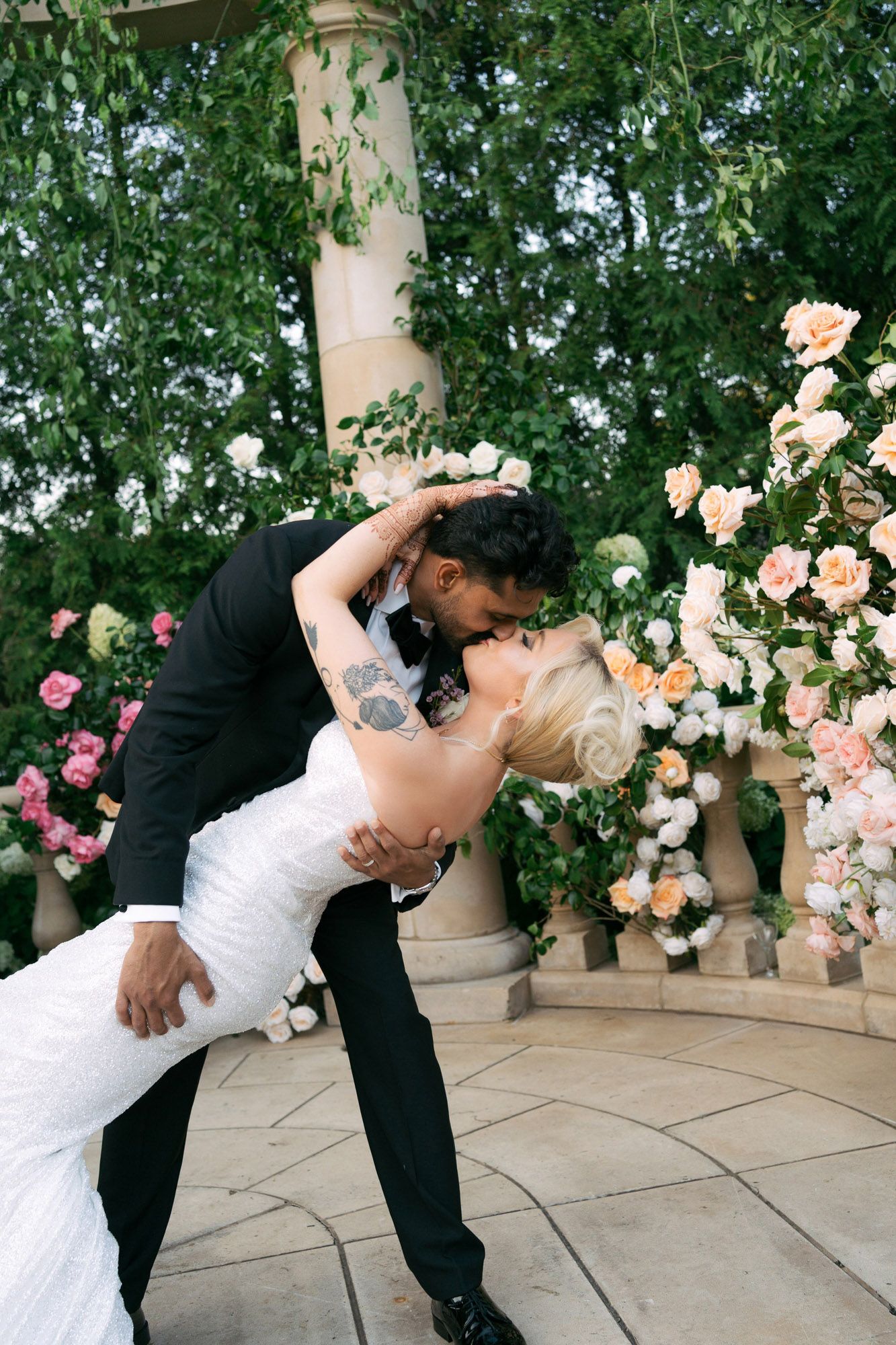 A romantic wedding couple dips and kisses in front of a floral arch with pink and peach roses in an outdoor setting.