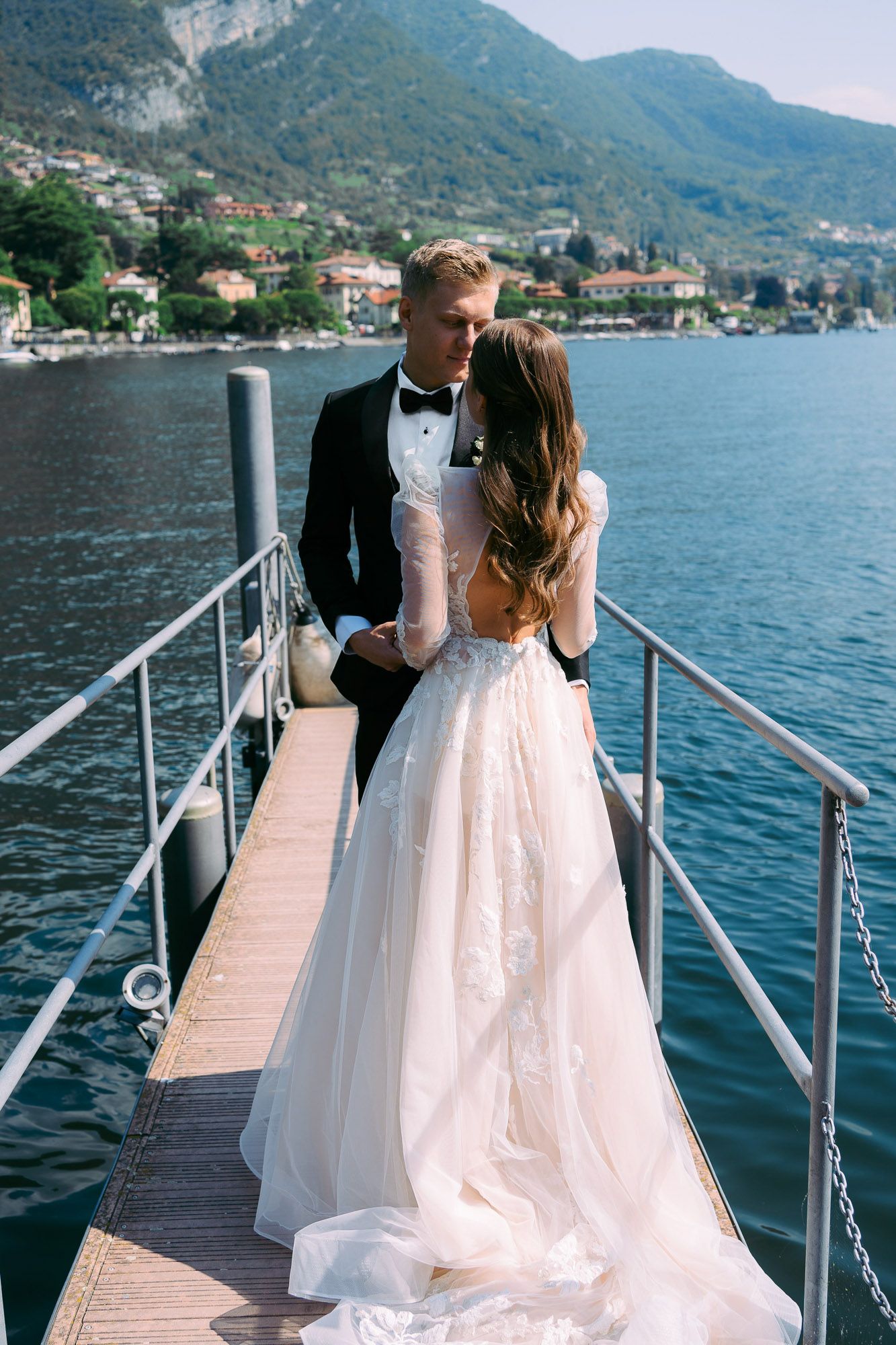 A couple in formal wedding attire stands on a wooden dock overlooking Lake Como with mountains in the background.