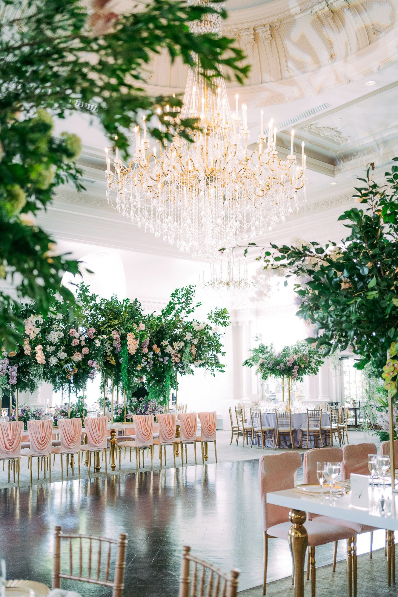 Elegant wedding reception venue with crystal chandeliers and lush greenery decorations in a bright white ballroom.