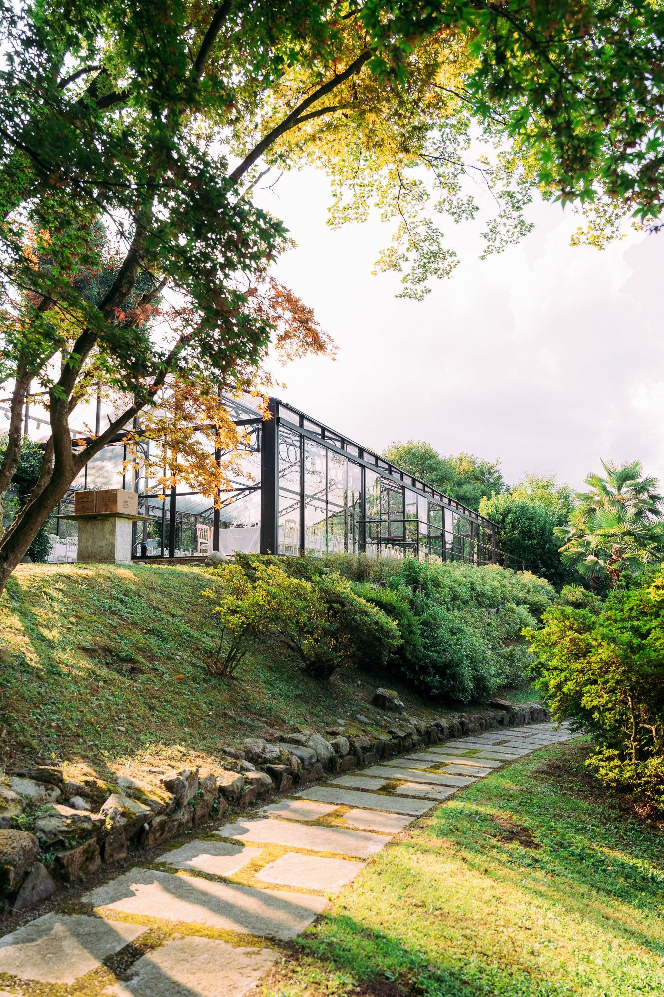 Stone pathway winding through a lush garden with glass greenhouse structure and autumn trees casting golden light.