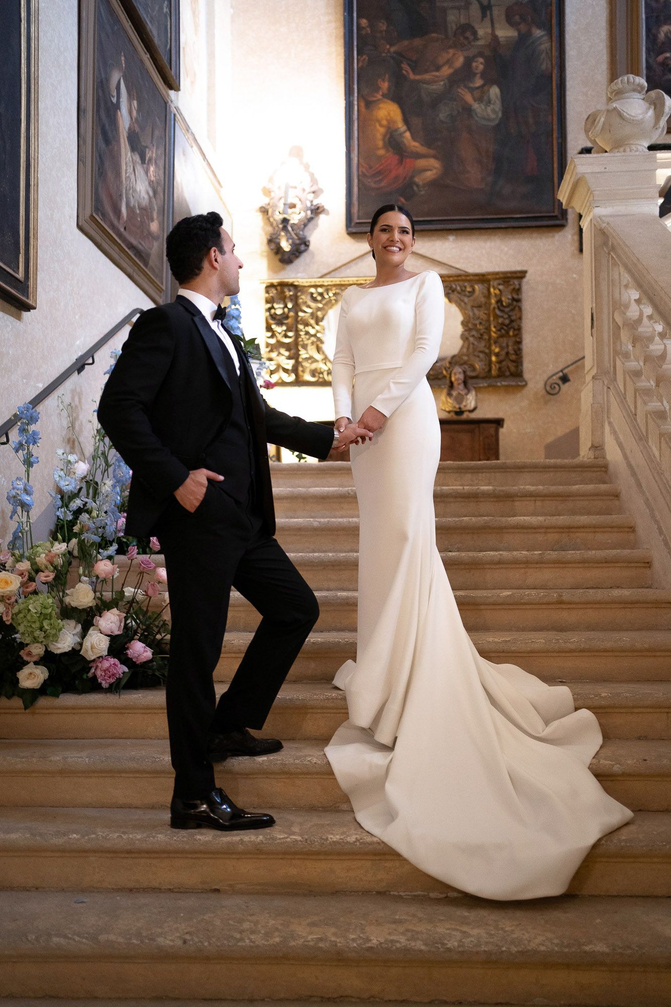 Elegant wedding couple posing on grand marble staircase adorned with floral arrangements.