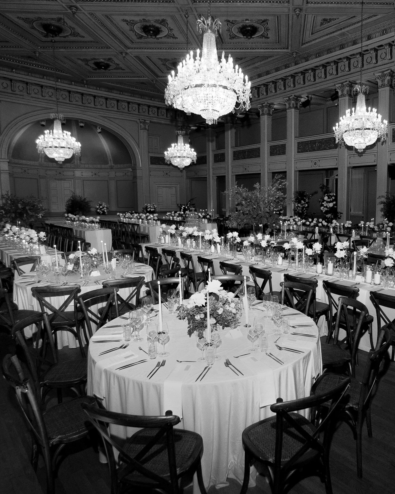 Elegant ballroom setup with round white tables, crystal chandeliers, and dark crossback chairs in a classic venue.