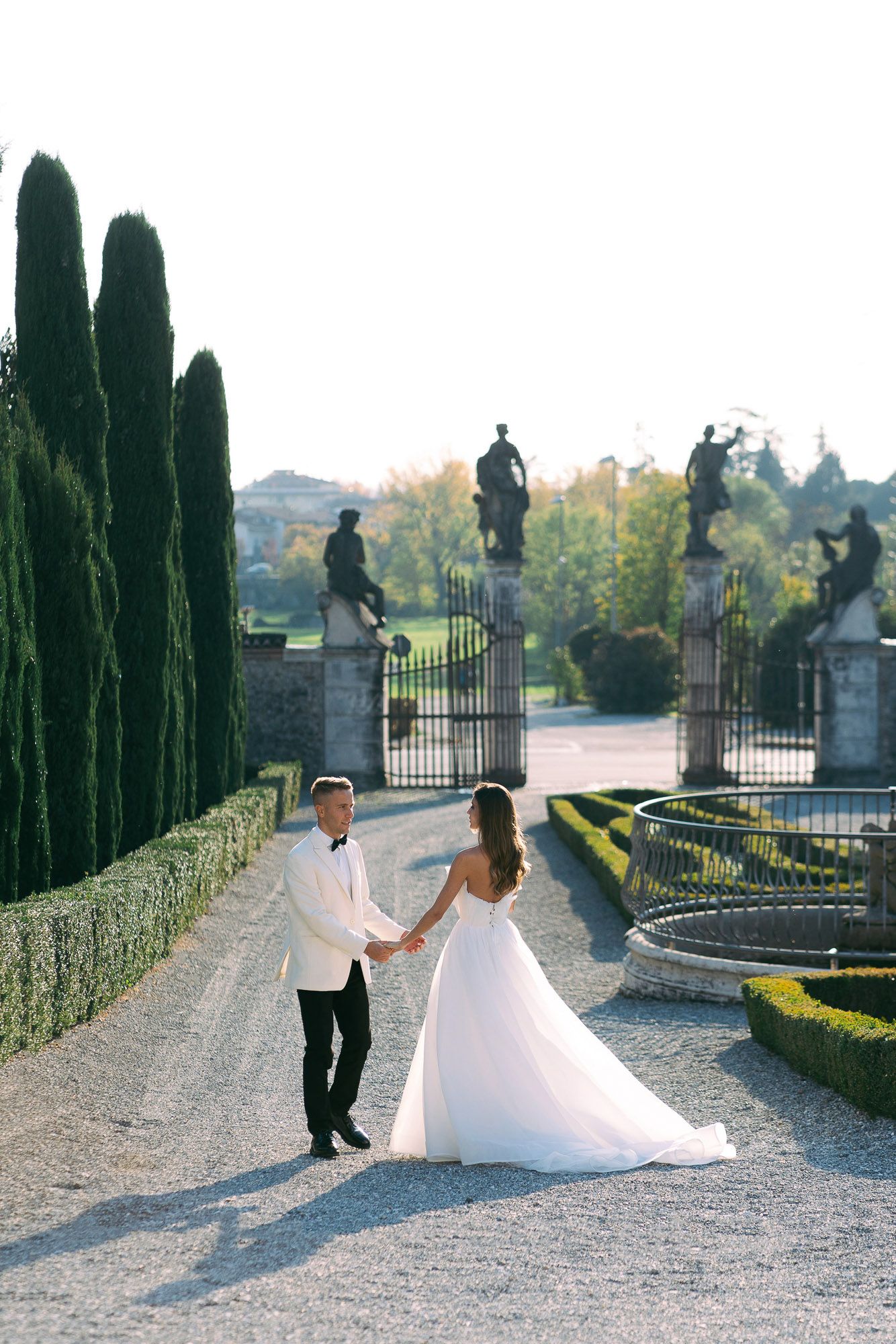 A romantic couple dances together on a gravel path lined with tall cypress trees and elegant stone gates at sunset.
