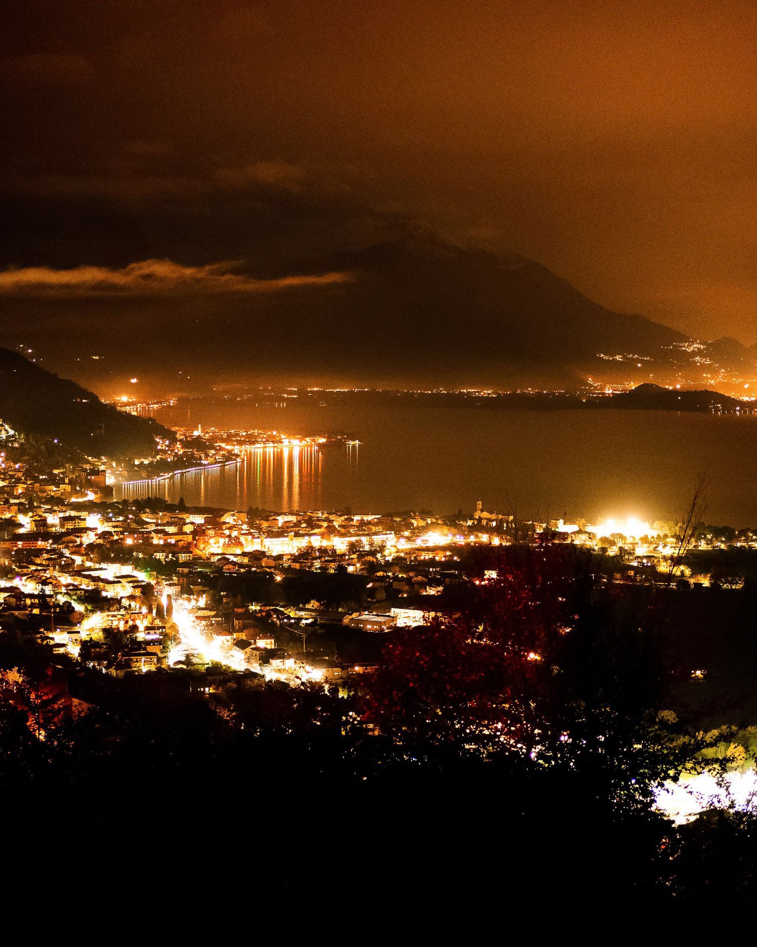Panoramic nighttime cityscape with twinkling lights along a mountainous coastline reflecting on dark waters.