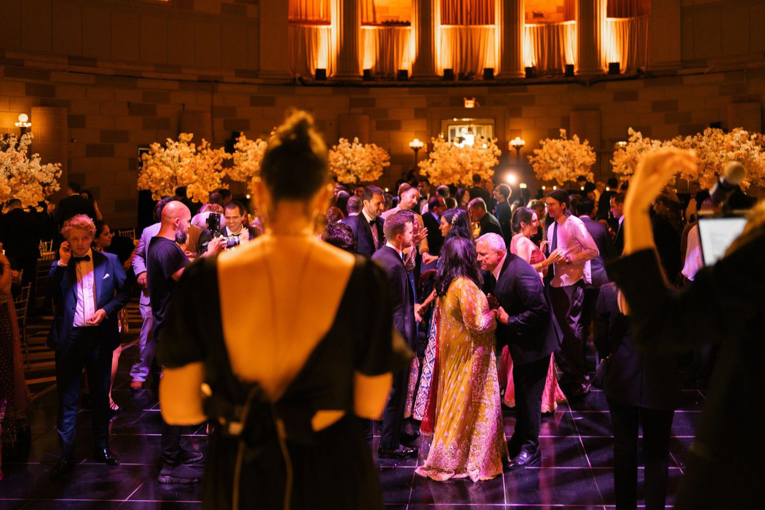 Wedding reception guests dance and celebrate under warm lighting with floral decorations in an elegant ballroom setting.