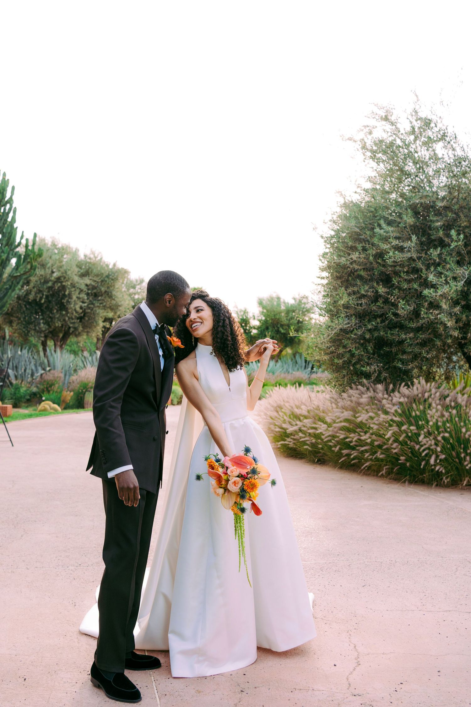 Bride and groom share intimate moment on garden pathway with pink and orange flowers.