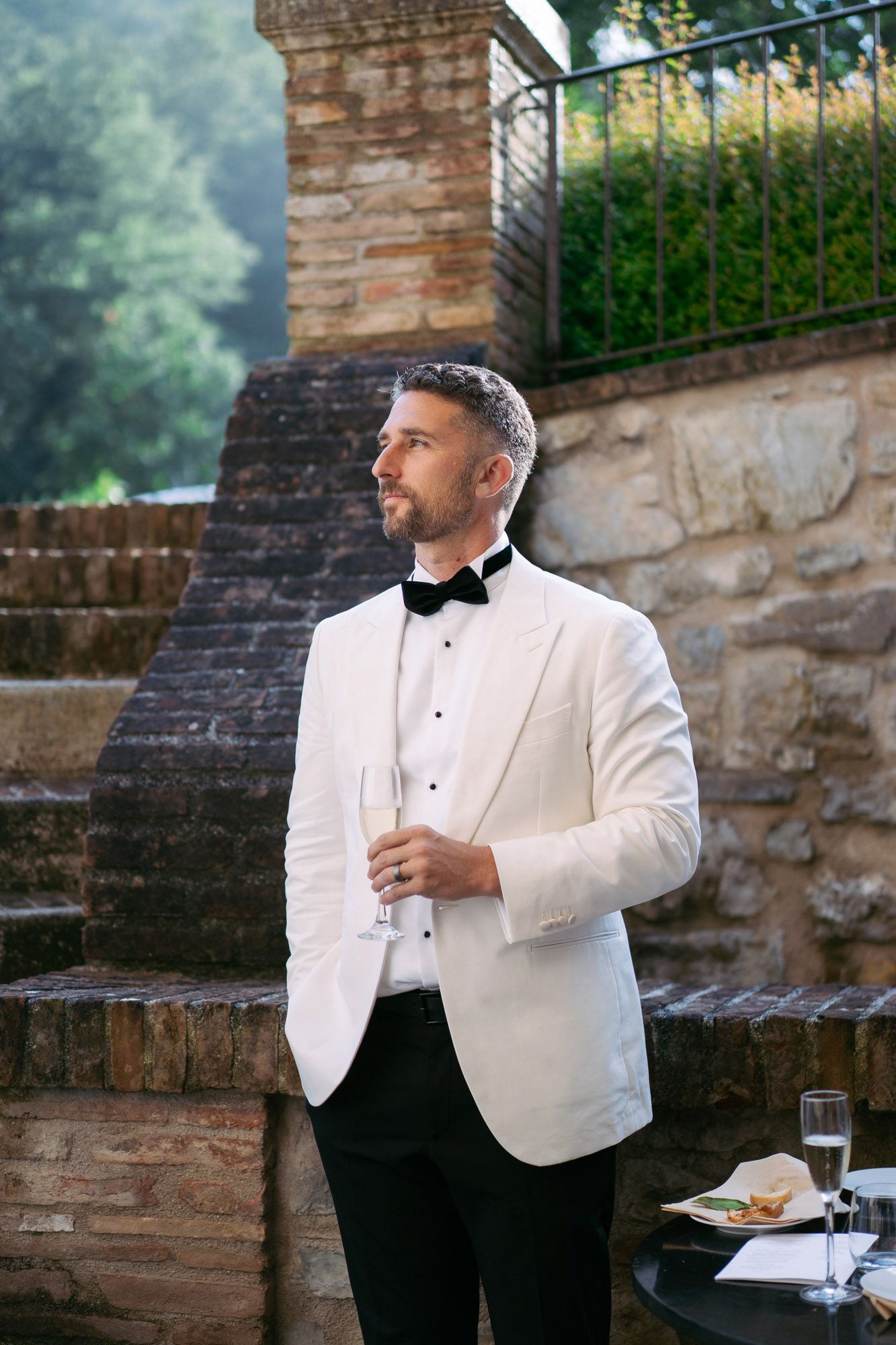 A groom in a white dinner jacket and black bow tie stands beside a stone wall during an outdoor wedding.