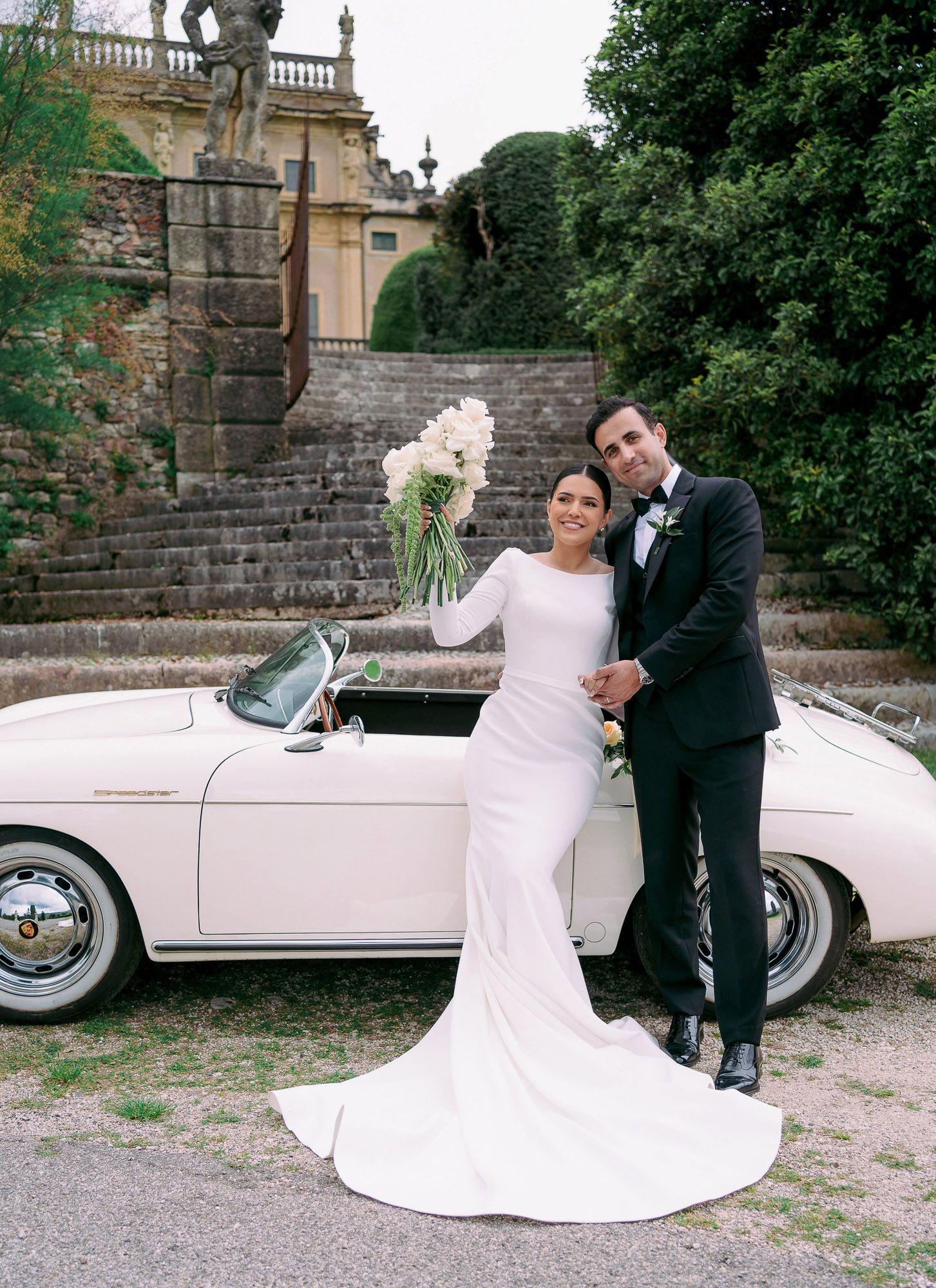 A couple in wedding attire poses elegantly beside a classic white vintage car in a garden setting.