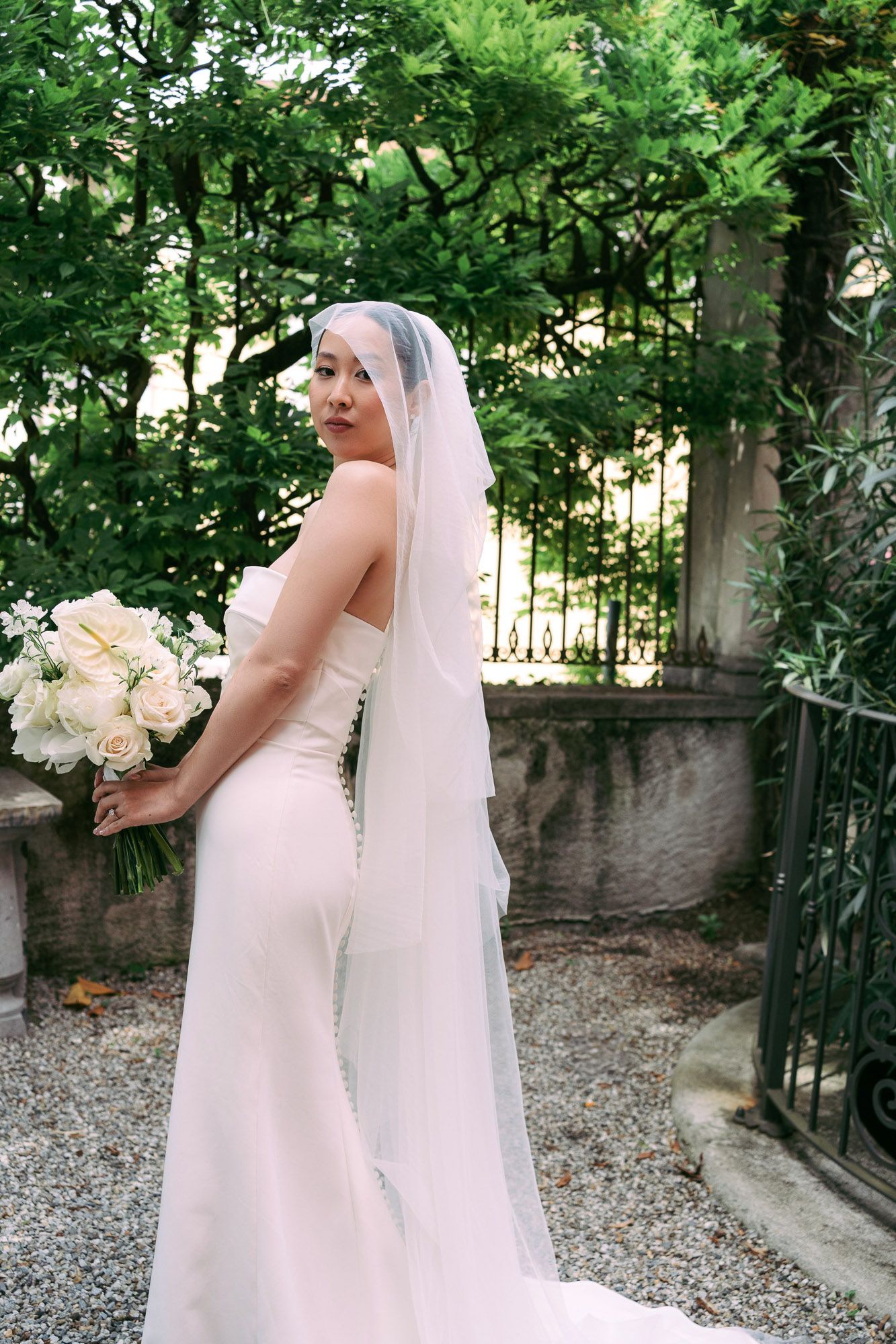 A bride in a white wedding dress and veil holds a bouquet of white flowers while standing near a stone wall and garden gate.