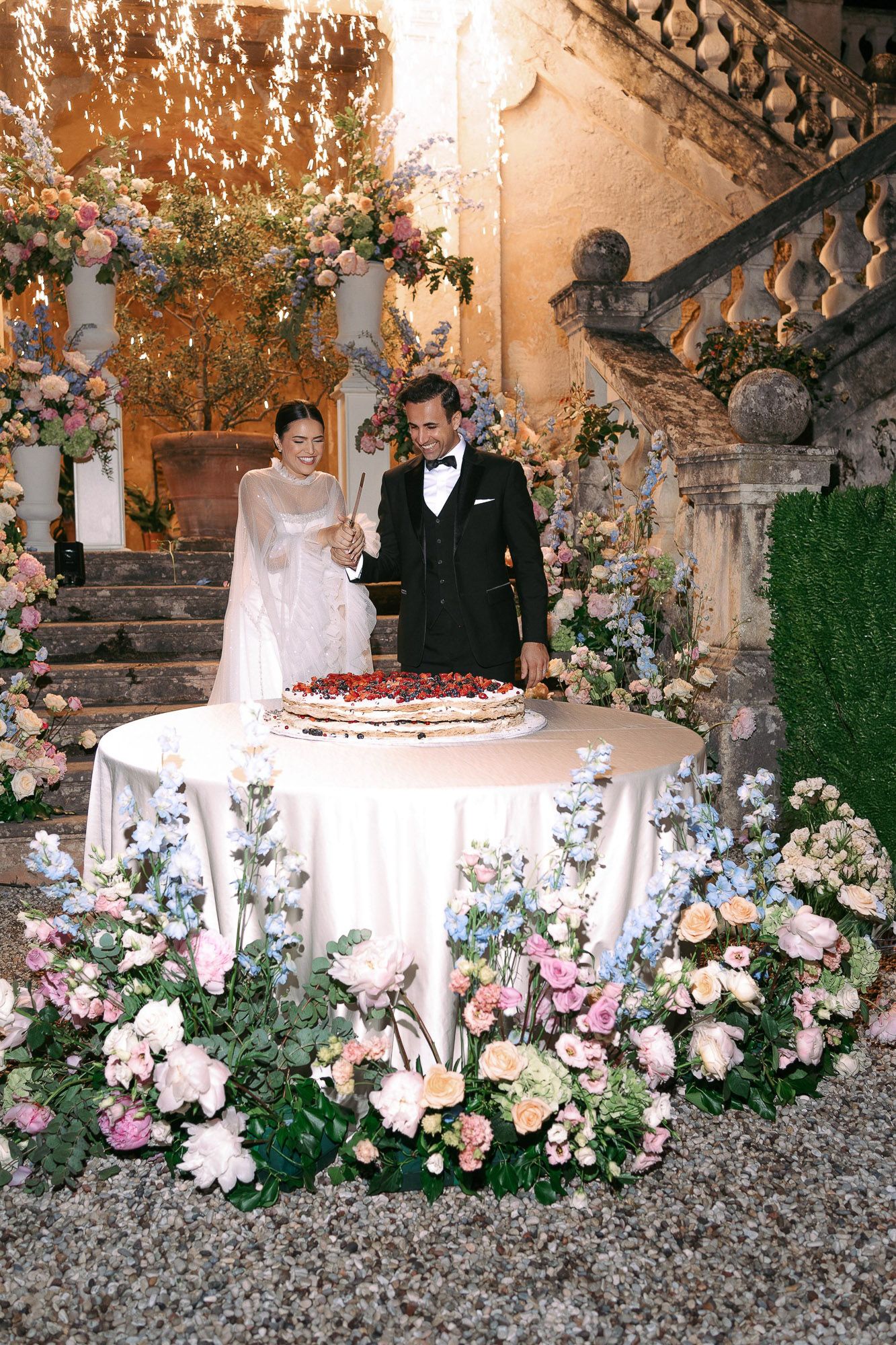 A romantic wedding cake cutting moment at an ornate stone staircase decorated with lush floral arrangements and greenery.
