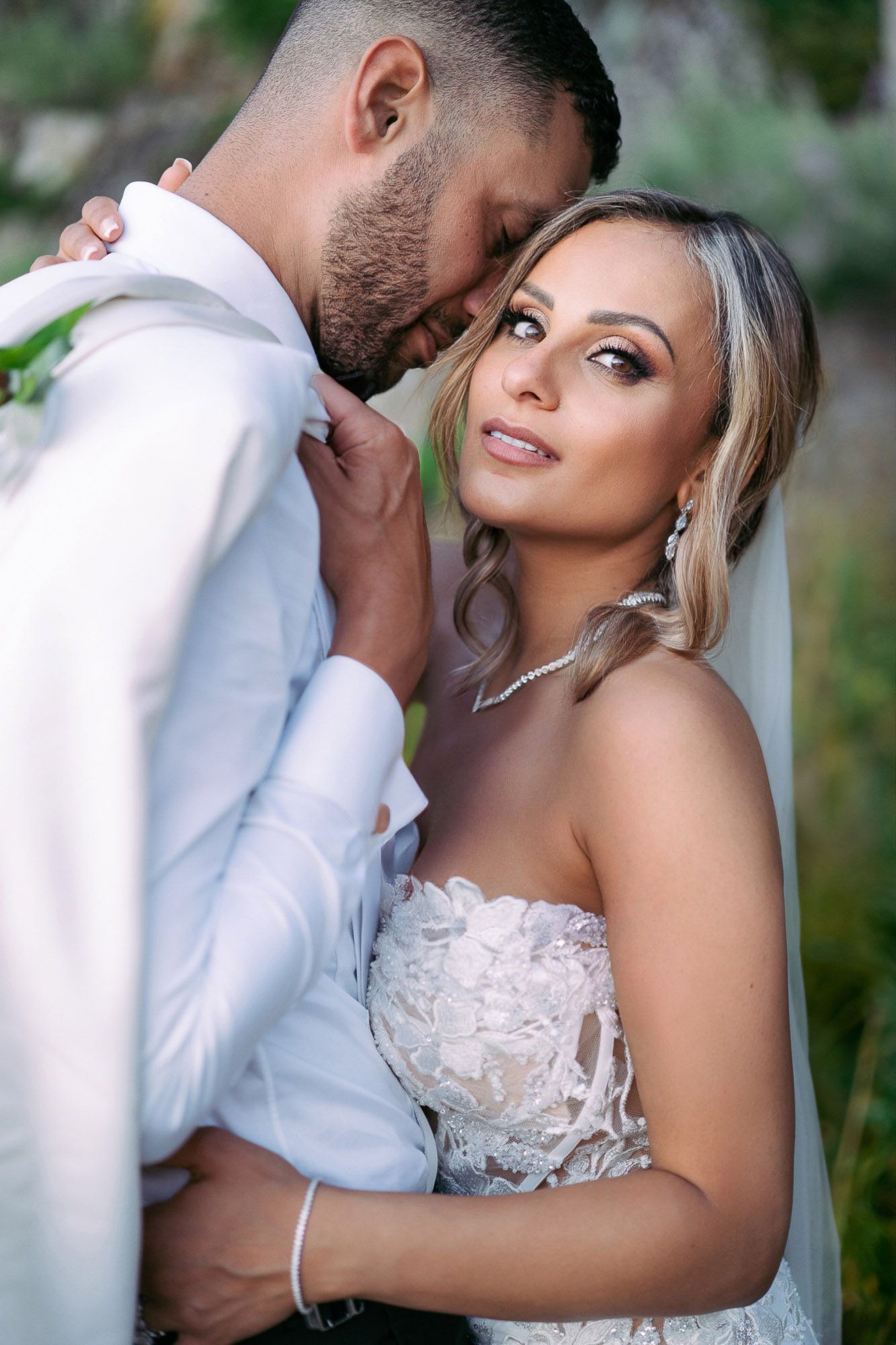A romantic wedding couple embraces in an outdoor garden setting wearing white formal attire.