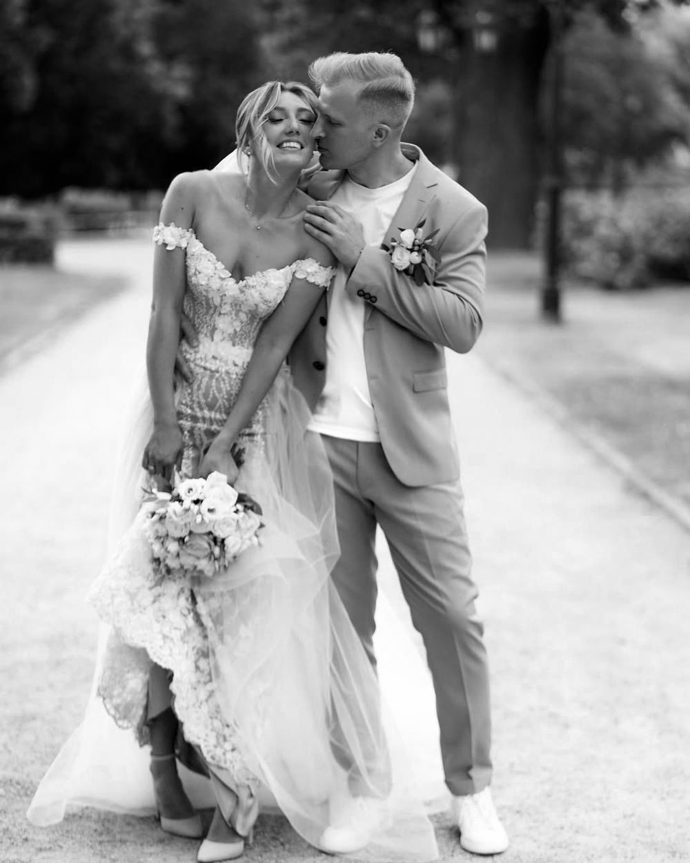 A joyful black and white wedding photo of a couple laughing together on a garden path with the bride holding a floral bouquet.