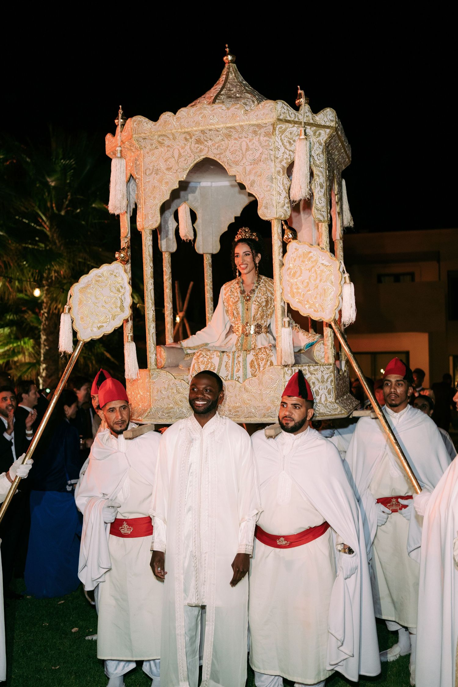 Traditional religious procession with people carrying ornate canopy structures while wearing white robes and red headwear.