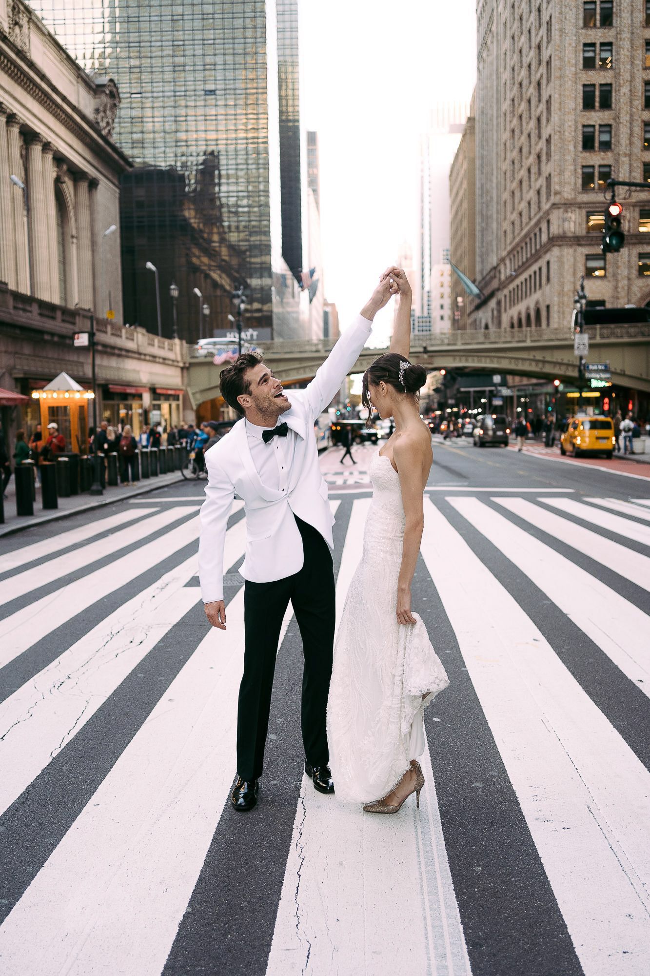 A couple performs dance lifts and spins on a zebra crossing in front of urban skyscrapers.