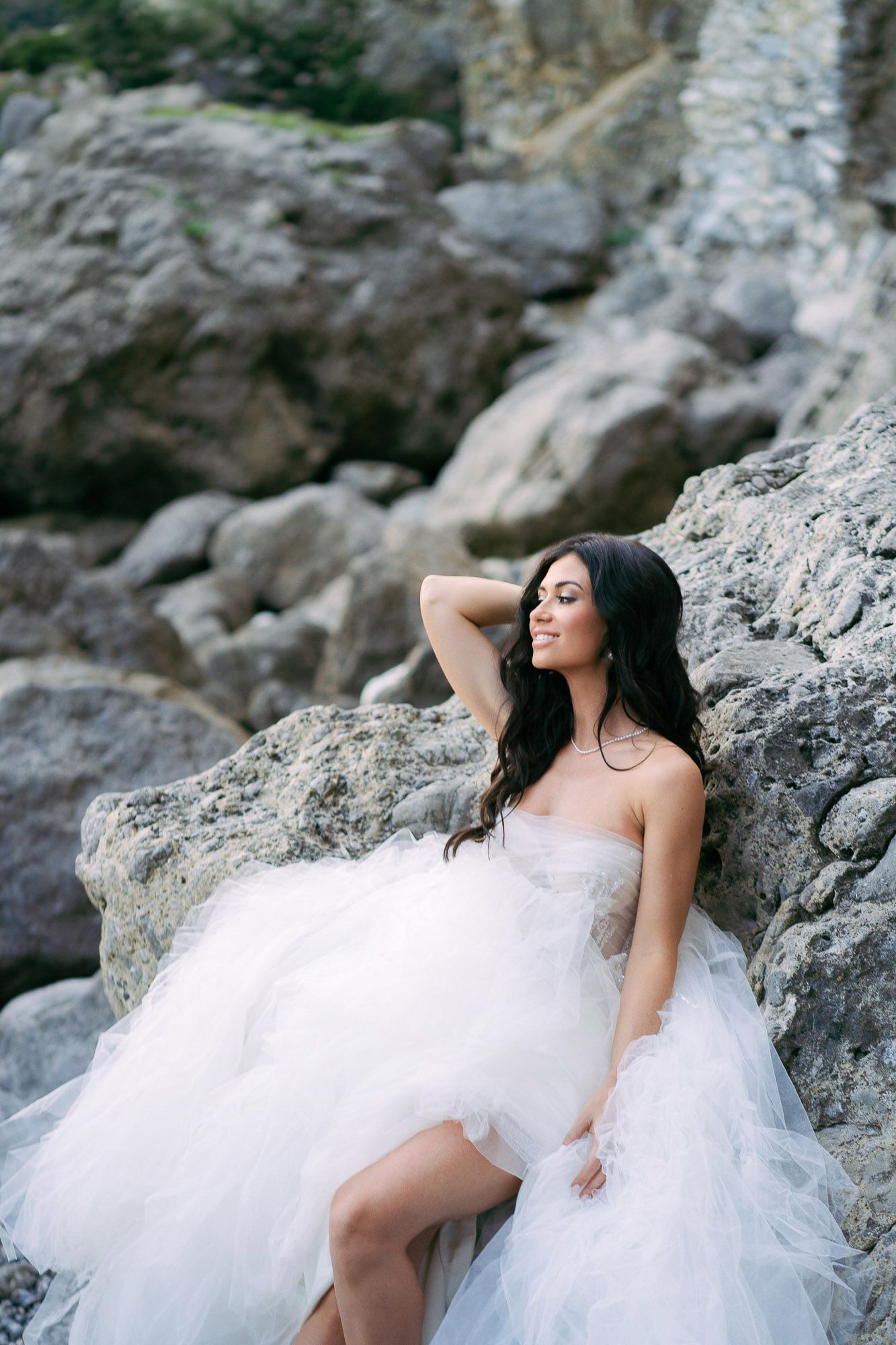 A bride in a flowing white tulle gown poses gracefully on coastal rocks during a dramatic seaside photoshoot.