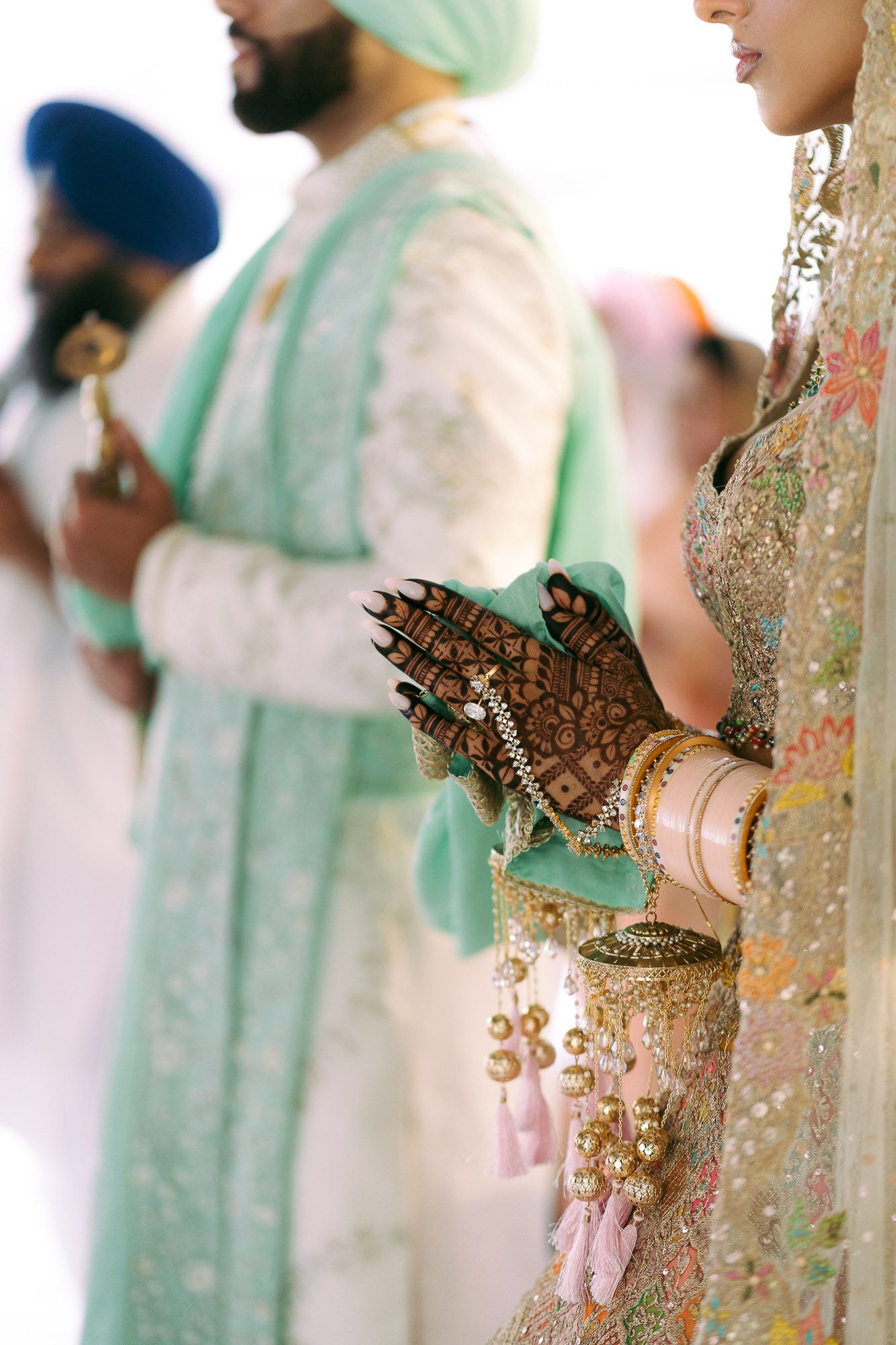 Close-up of ornate henna designs and traditional bridal jewelry on hands against mint green fabric.