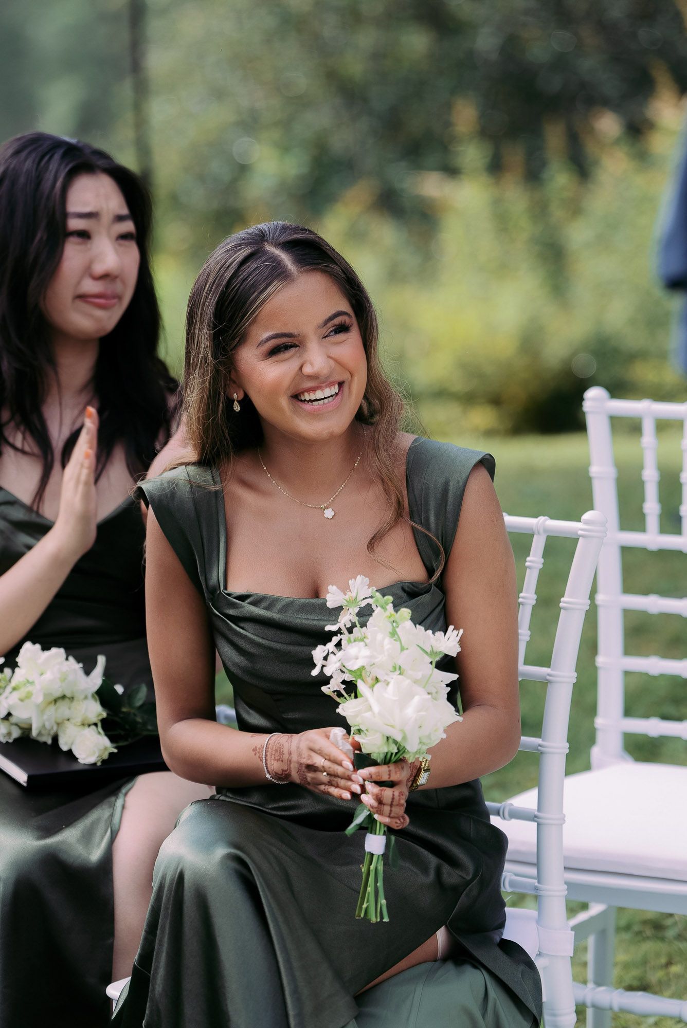 Bridesmaids in dark green dresses hold white floral bouquets while sitting in white chairs at an outdoor wedding ceremony.