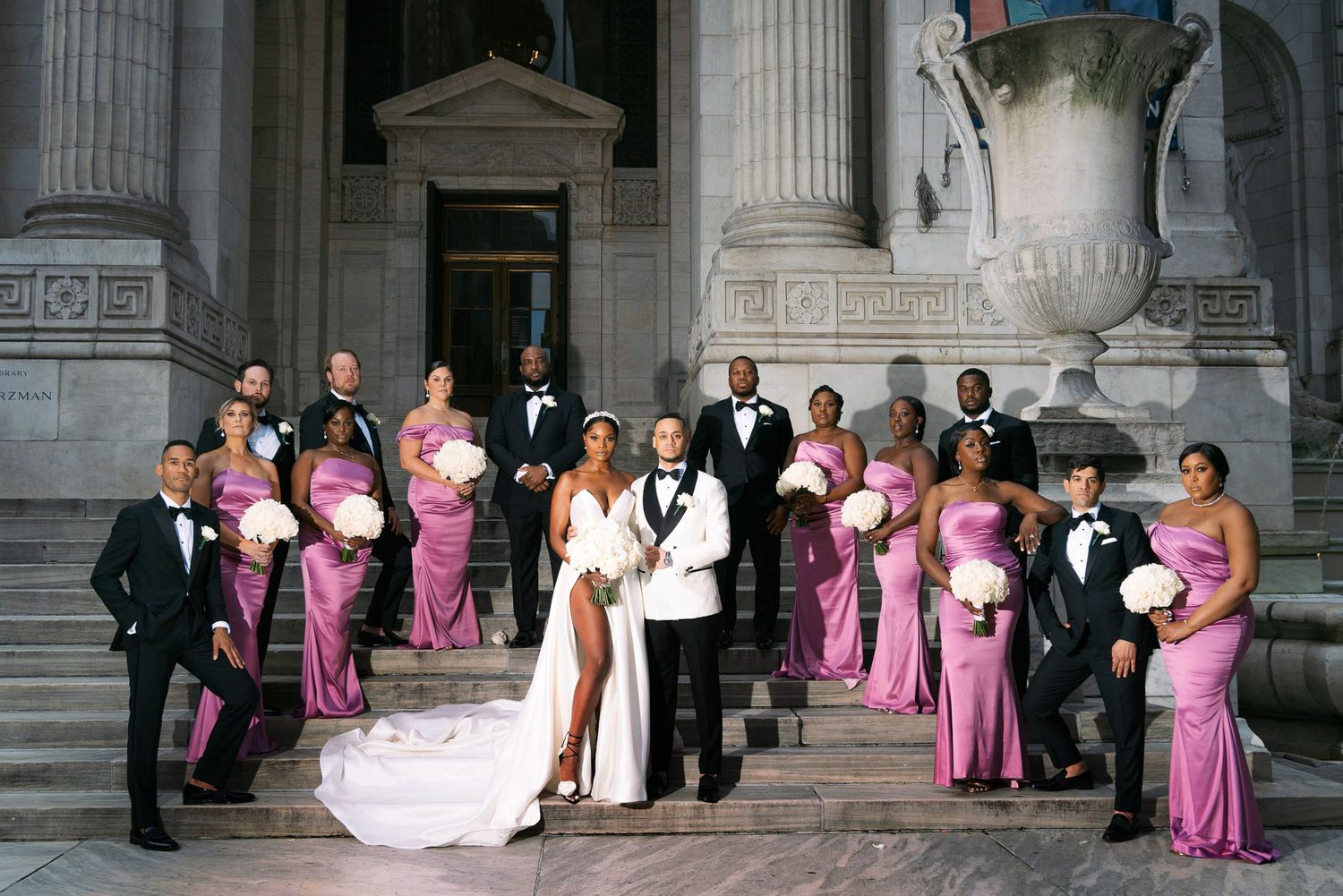 Wedding party poses on grand stone steps of historic building with classical columns in formal attire.