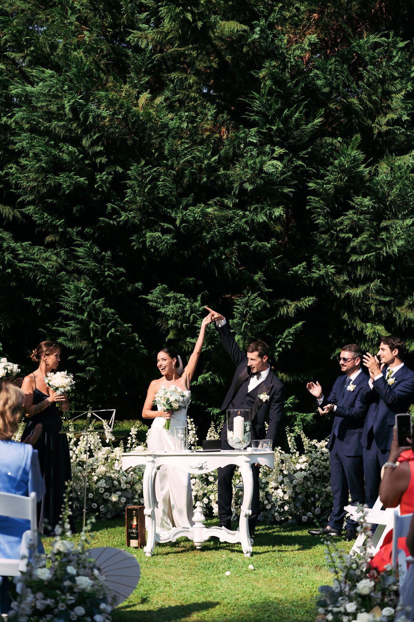 A wedding party celebrates outdoors in a garden setting with trees and greenery in the background.