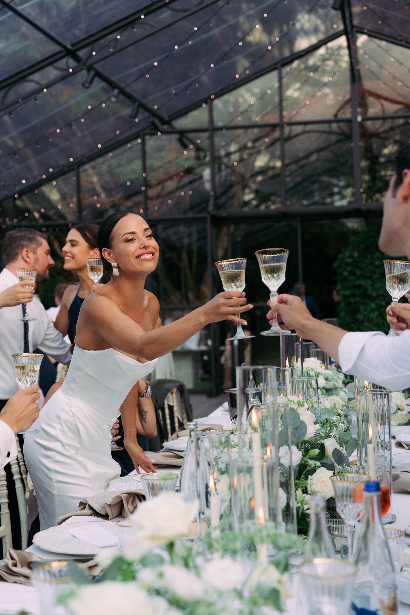 Wedding guests raise glasses in celebration during an elegant outdoor reception under a glass-roofed venue.