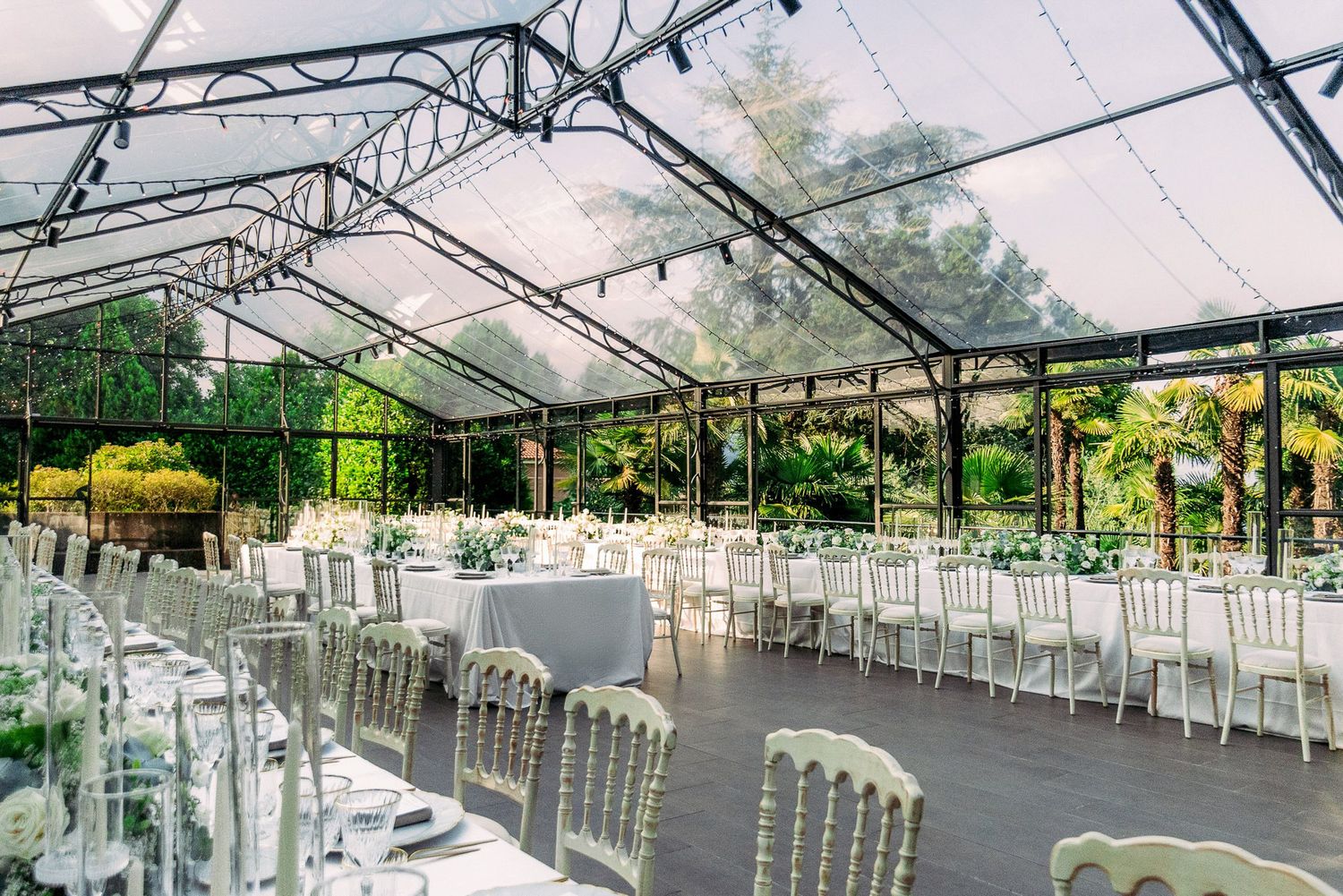 White wedding reception chairs arranged in rows under a clear tent canopy with hanging string lights and chandeliers.