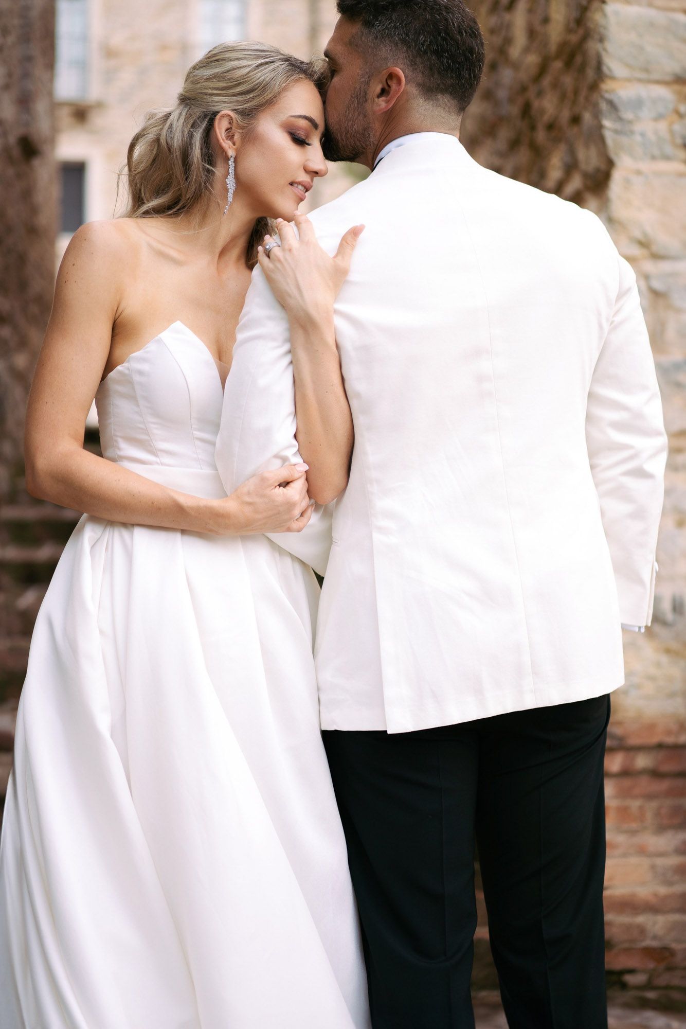 A romantic couple in white attire embracing against a stone wall in a series of intimate wedding photos.