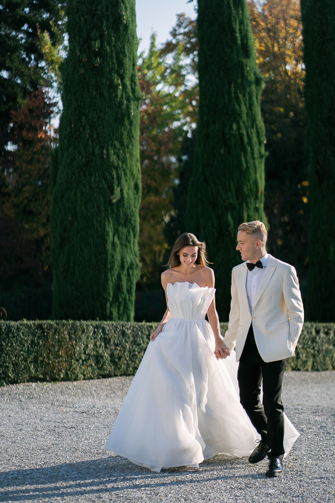 A couple in wedding attire walks hand in hand along a cypress tree-lined path in an elegant outdoor setting.