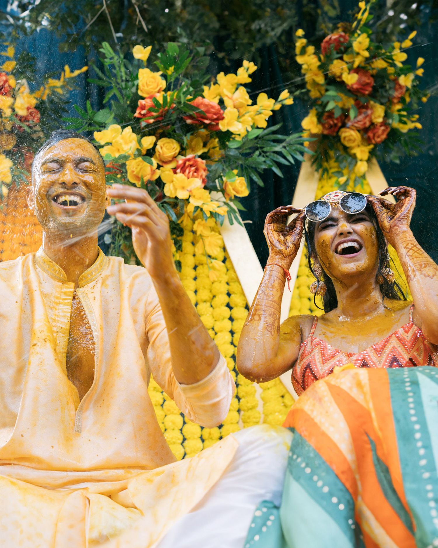 Group of people wearing bright yellow flower crowns laugh joyfully while dressed in light colored outfits.