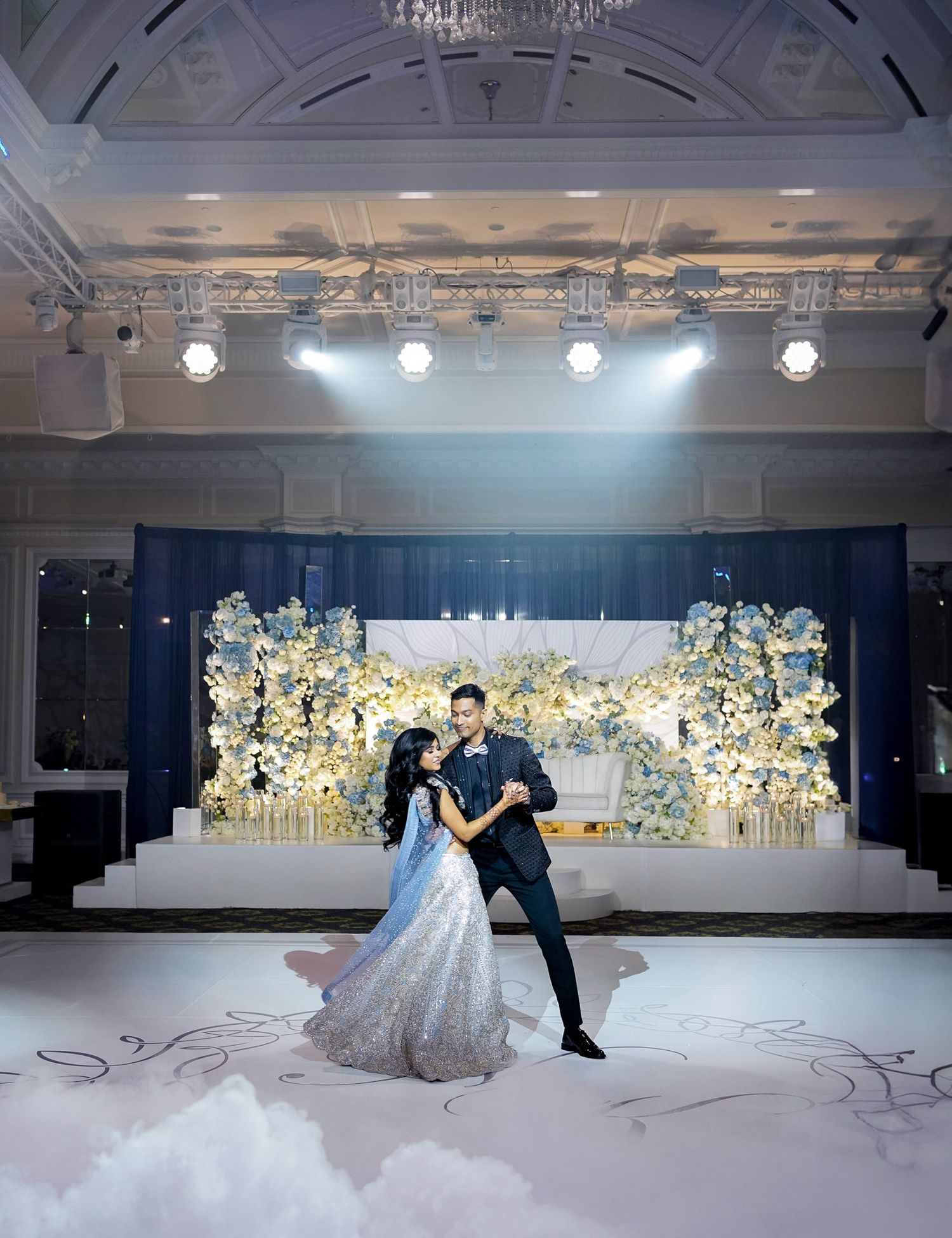 A couple dances in an elegant ballroom with white floral decorations and chandeliers creating a romantic atmosphere.