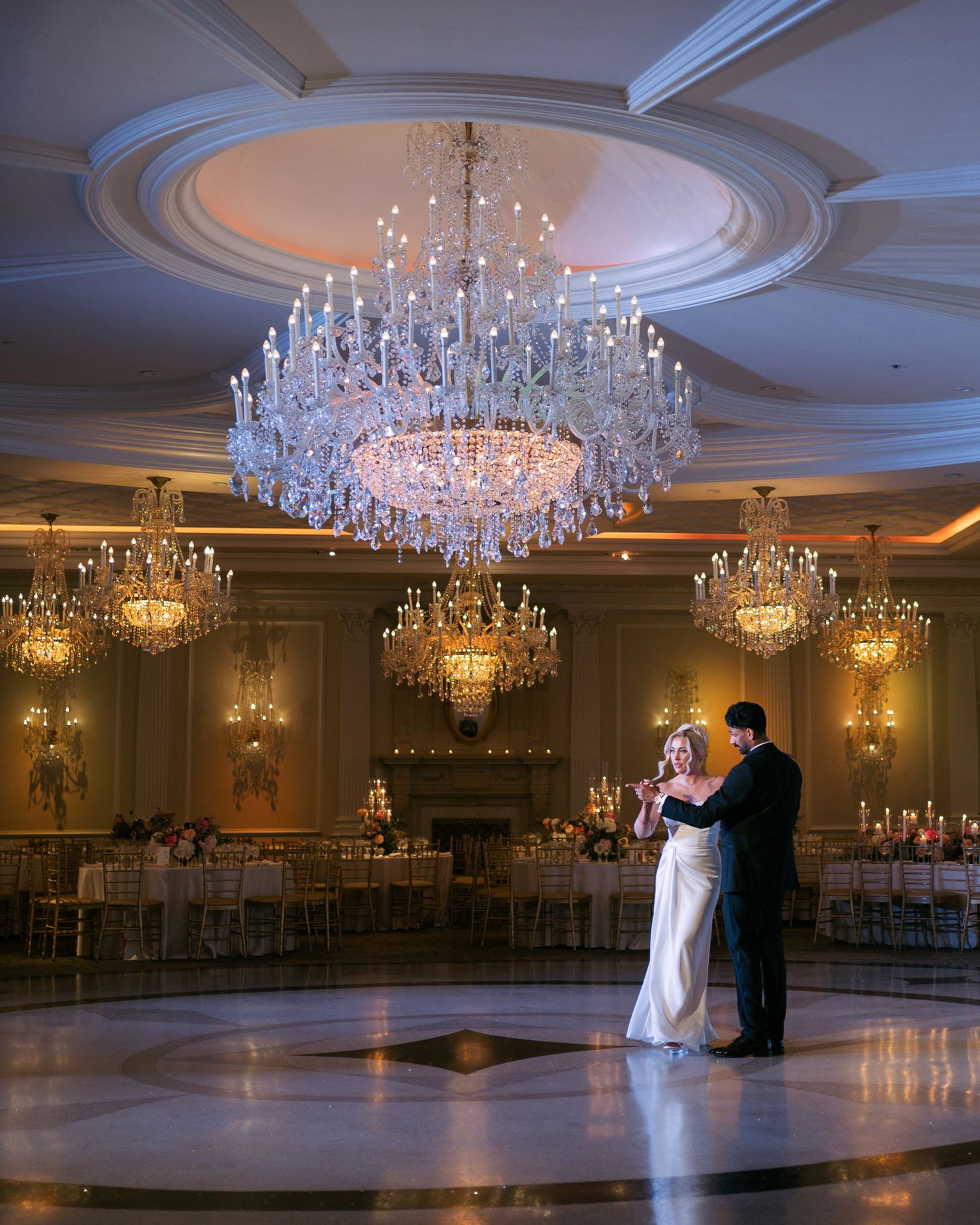 Wide view of an opulent ballroom with multiple crystal chandeliers and a couple dancing on the polished floor.