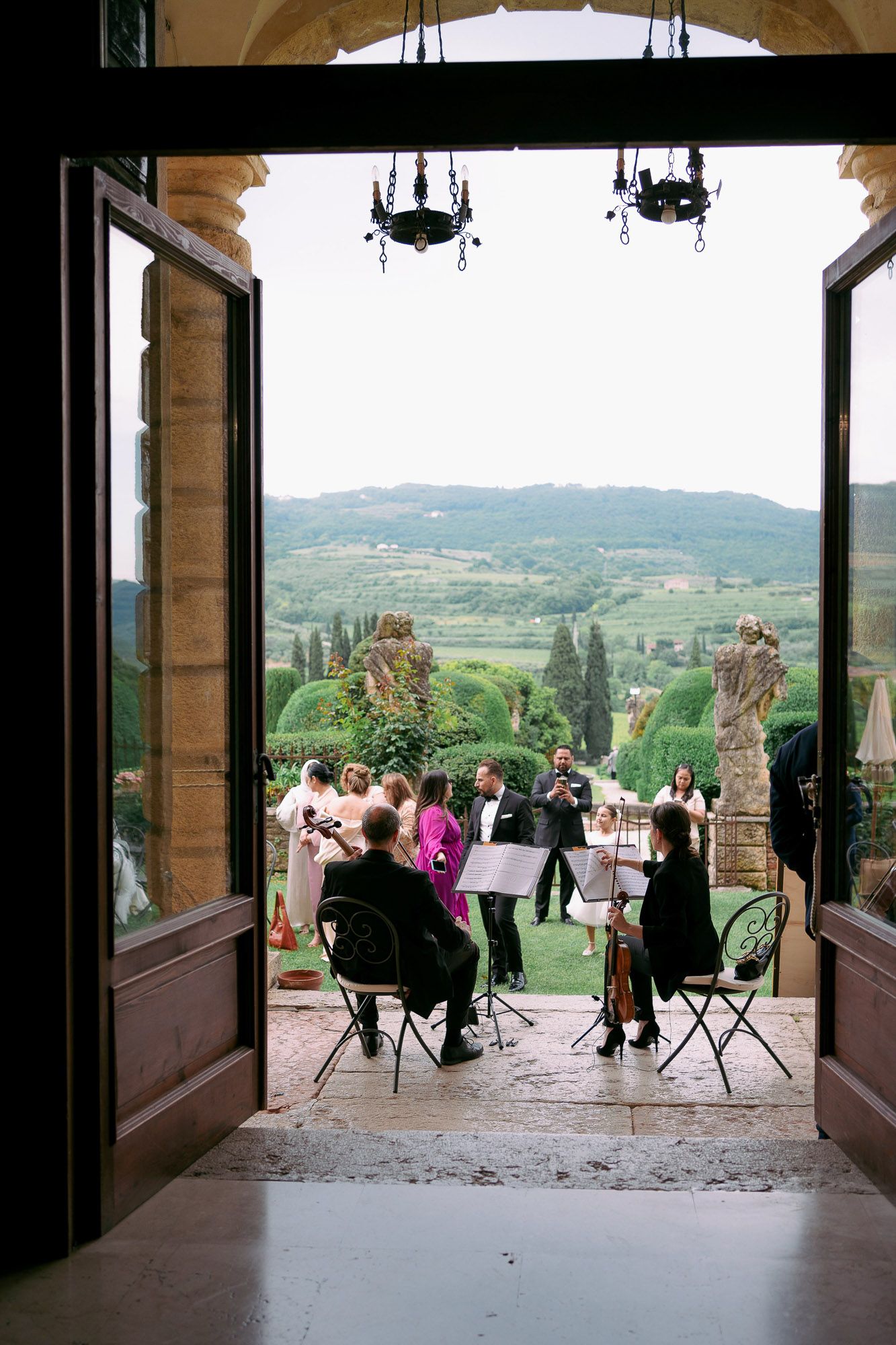 A group of people sit on chairs at a Tuscan villa terrace overlooking scenic rolling hills and vineyards in Italy.