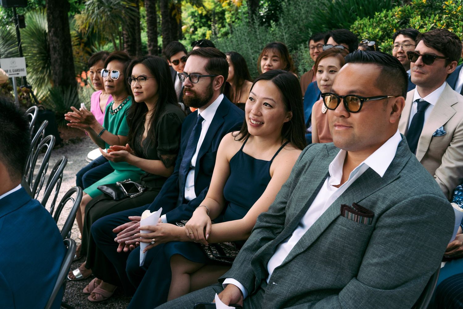 Wedding guests seated outdoors in formal attire watch a ceremony on a sunny afternoon.