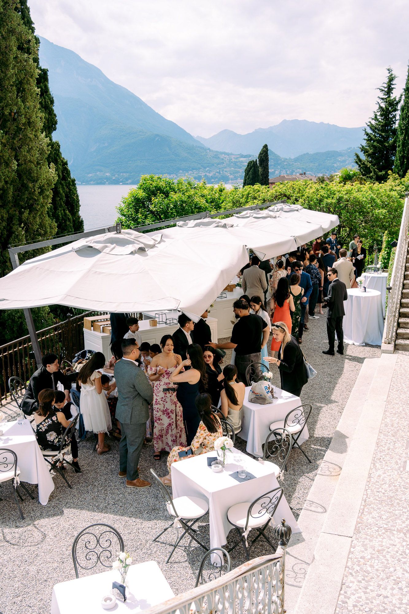 People gather under white canopy tents on a scenic terrace overlooking mountains and a lake in Italy.