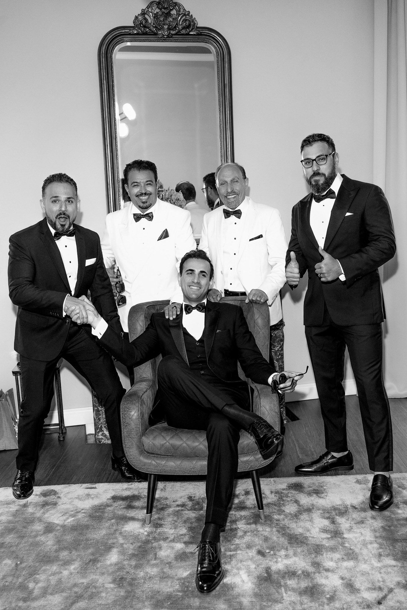 Five men in formal black tuxedos pose elegantly around an ornate chair against a light-colored wall.