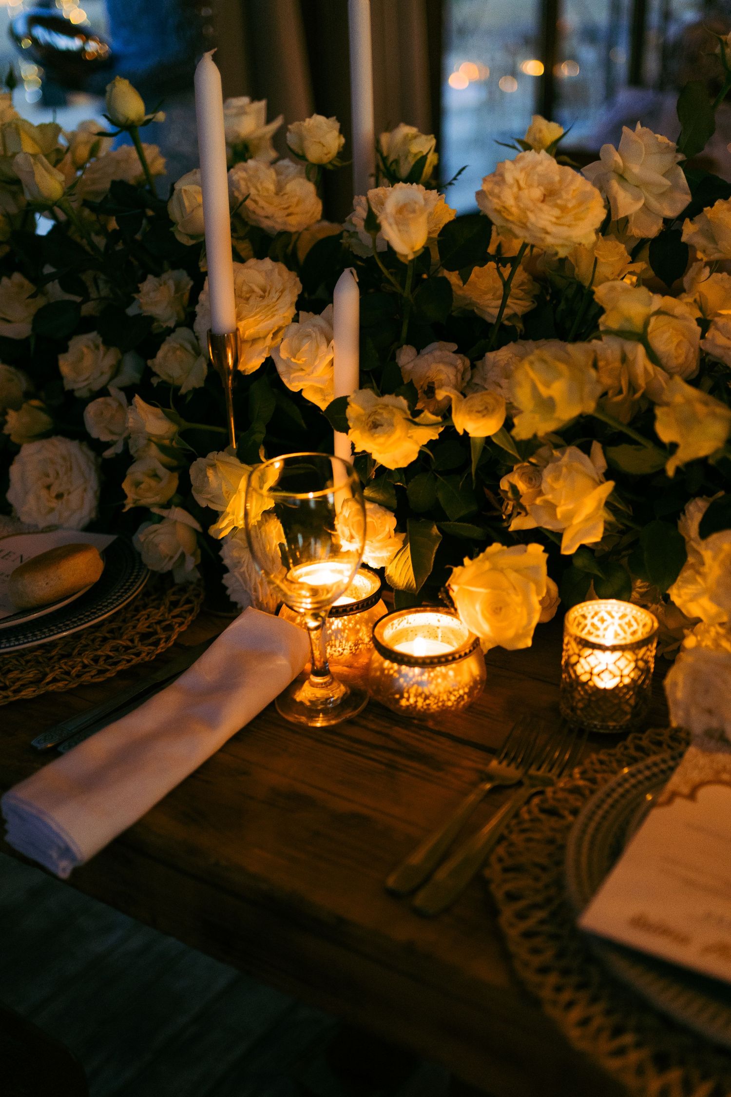 Romantic table setting with glowing candles in glass holders and yellow roses creating ambient lighting.