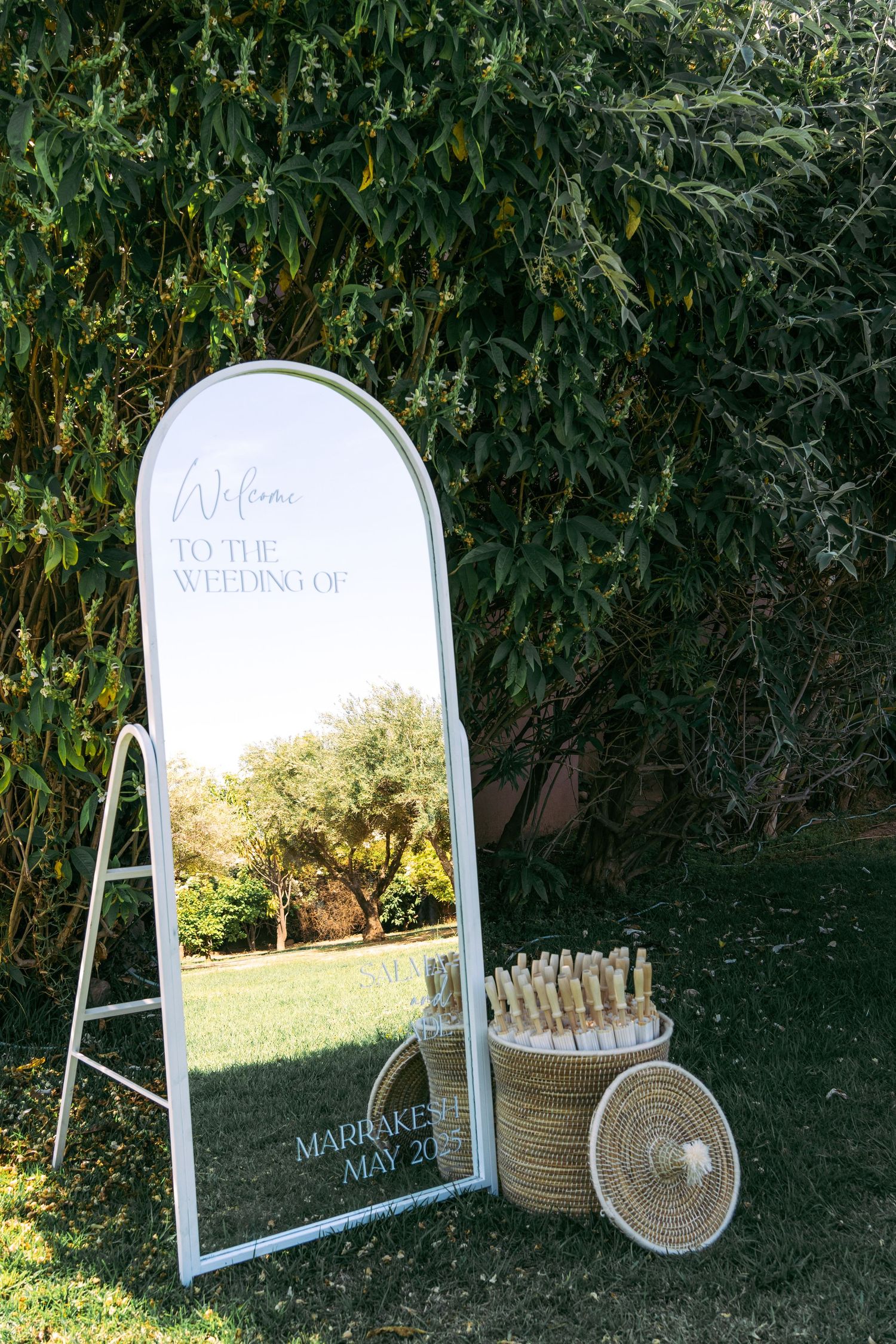 White decorative mirrors and display tables set up outdoors against dark foliage for a wedding event.