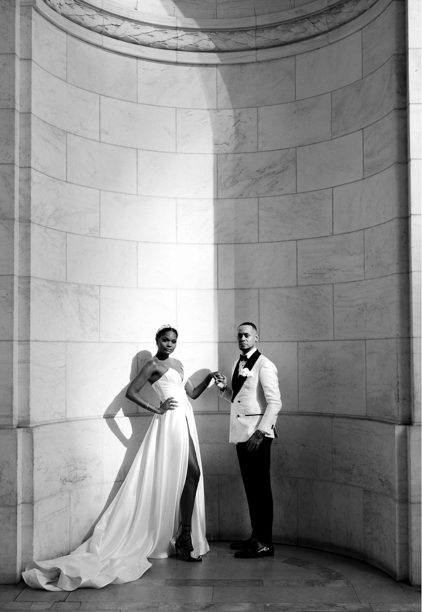A black and white wedding photo series showing a couple in formal attire posing against curved marble walls.