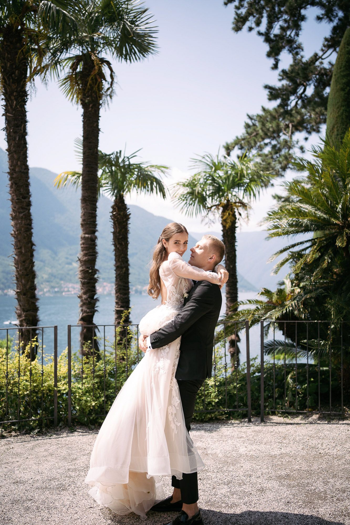 A romantic couple shares an intimate dance moment on a palm-lined terrace overlooking a scenic lake and mountains.