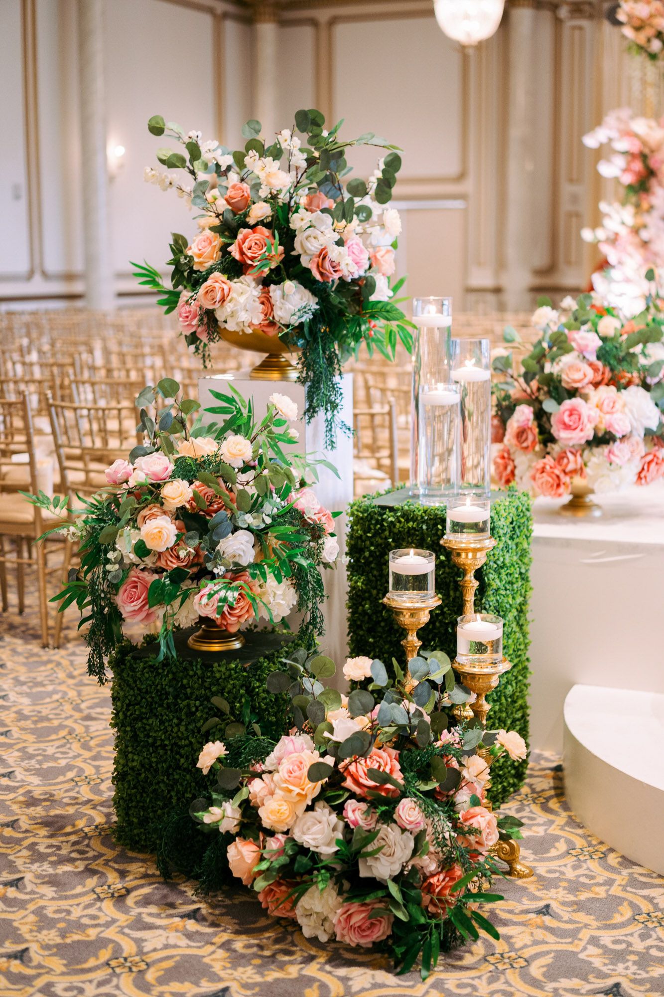 Pink roses and greenery line a wedding aisle with elegant floral arrangements on a patterned carpet.