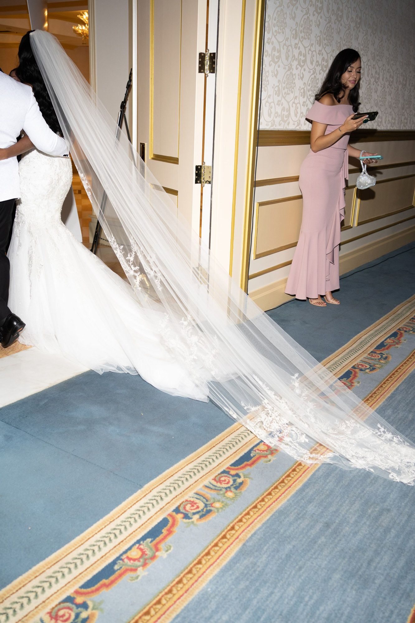 A flowing white wedding dress train trails elegantly across a patterned hotel hallway carpet.