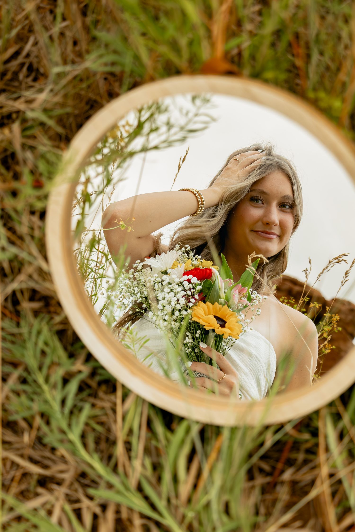 A circular mirror reflection in grass shows someone in white holding a colorful bouquet of flowers and baby's breath.