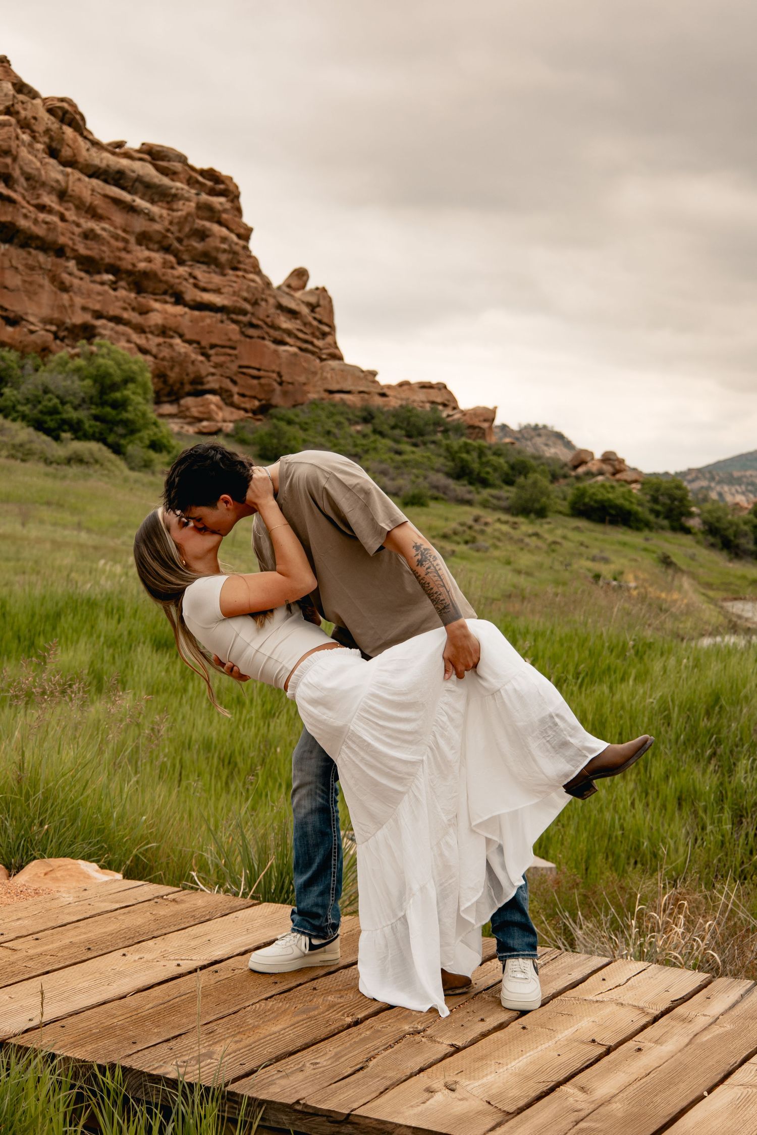 A romantic dip pose on a wooden deck against red rock formations and green grass with dramatic cloudy sky.
