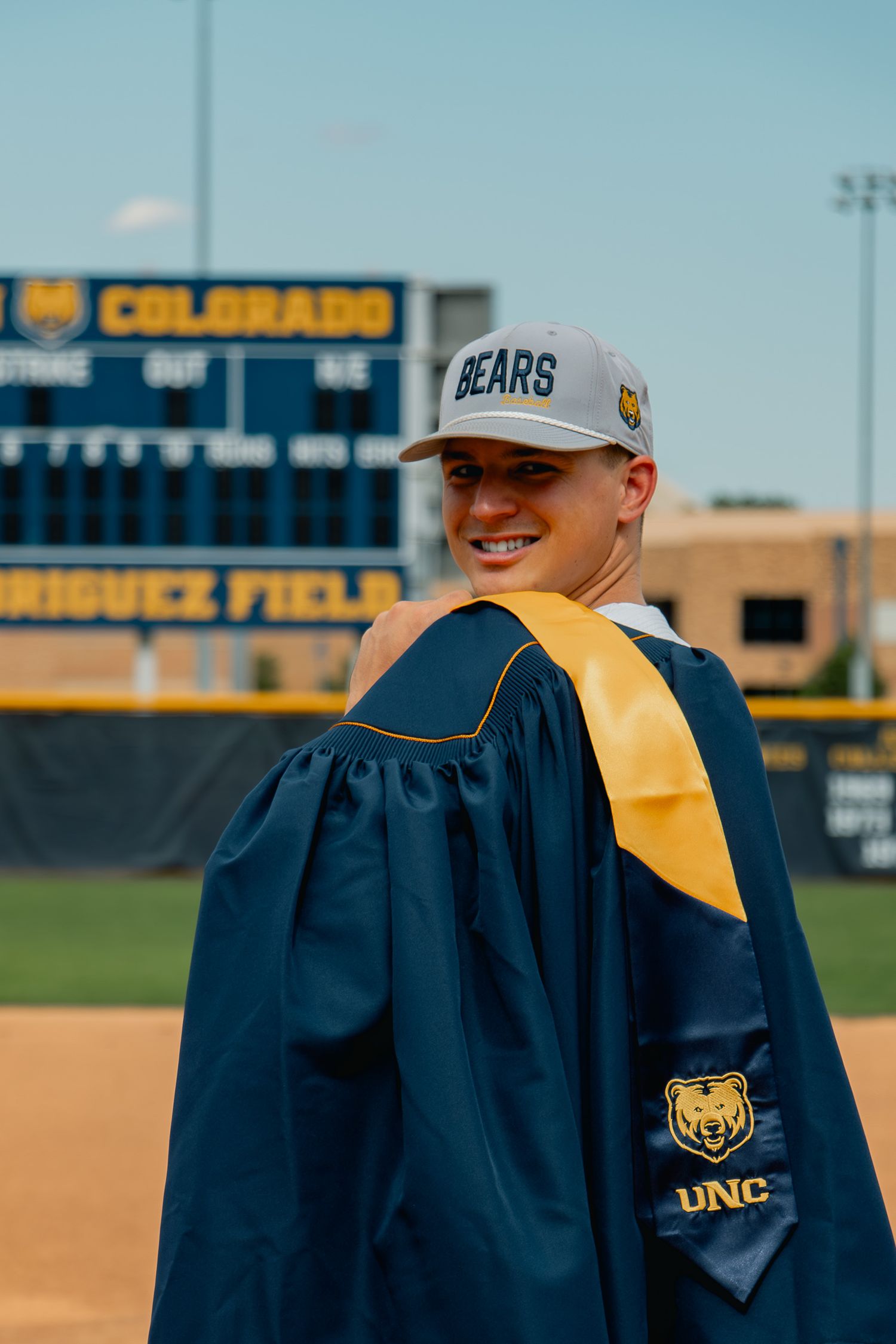 College graduate wearing blue and gold graduation cap and gown celebrates at baseball field with clear sky backdrop.