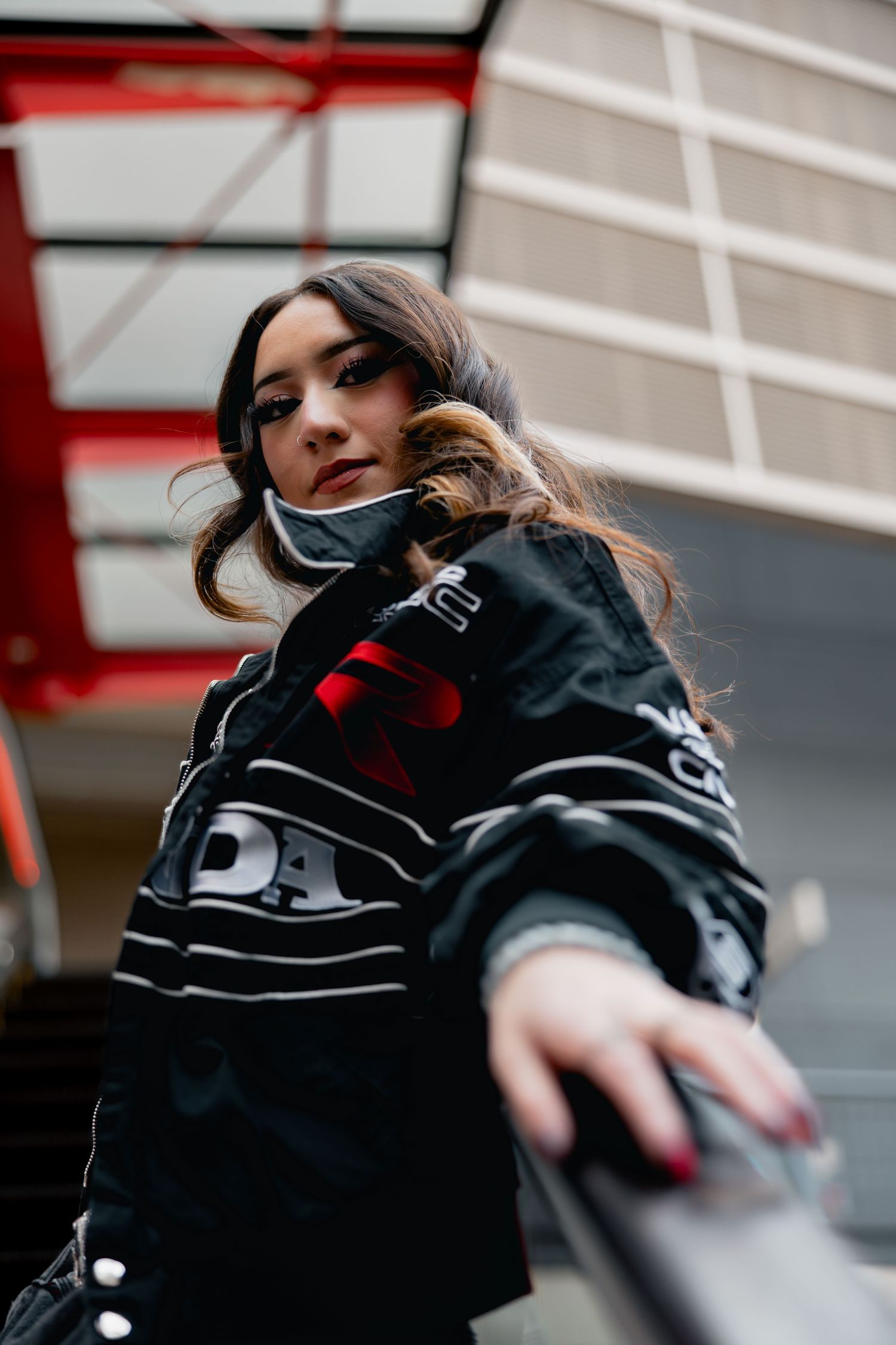 Young woman in black jacket leaning on railing, urban setting with red accents.