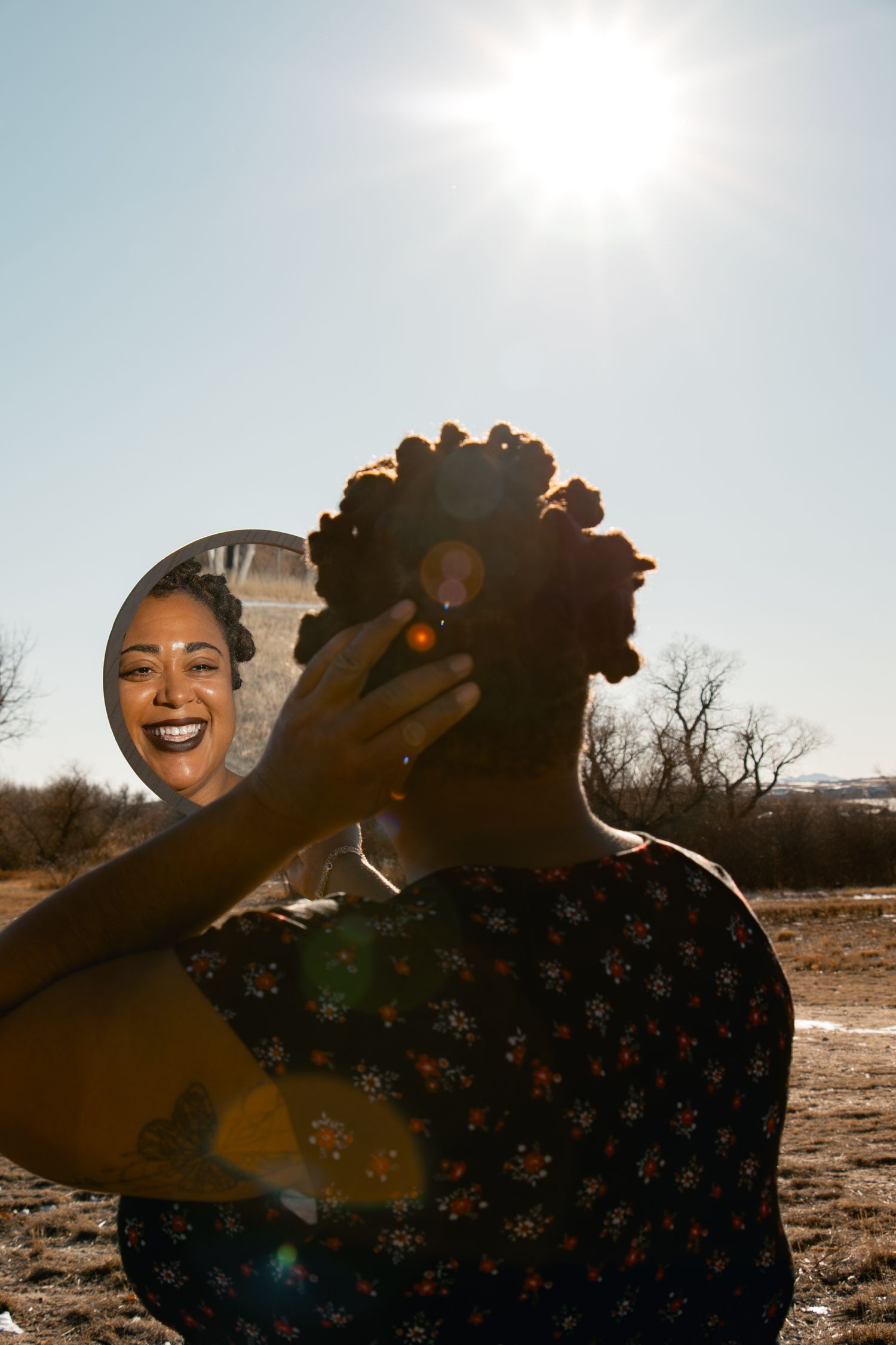 A smiling Black woman holds a mirror reflecting her joyful face outdoors in bright sunlight, wearing a floral dress.